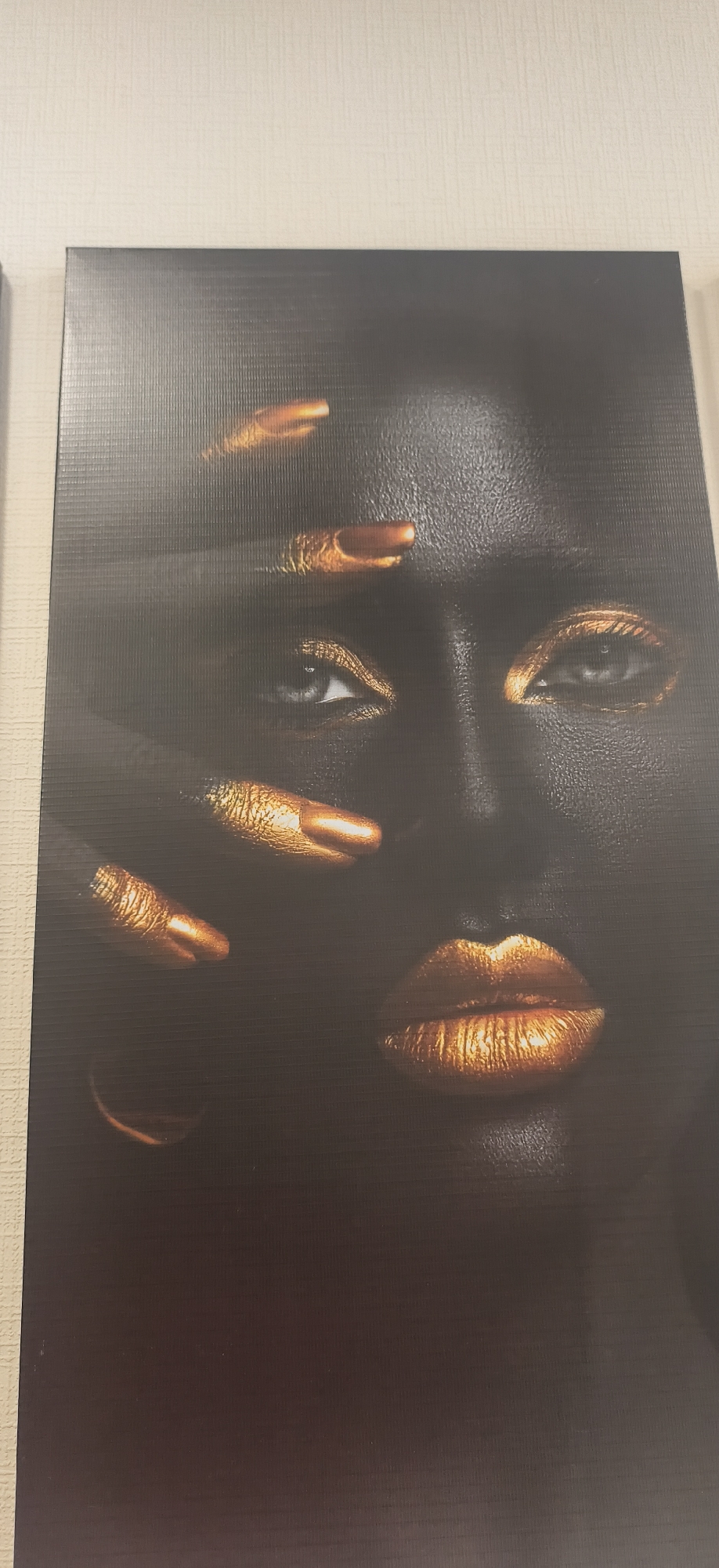 The image shows a close-up portrait of a woman with dark skin and golden accents. Her face is mostly shadowed, but her eyes, lips, and fingernails have a metallic gold color. One hand is partially obscuring her face. The background is dark and indistinct. The location is Unknown, Unknown.