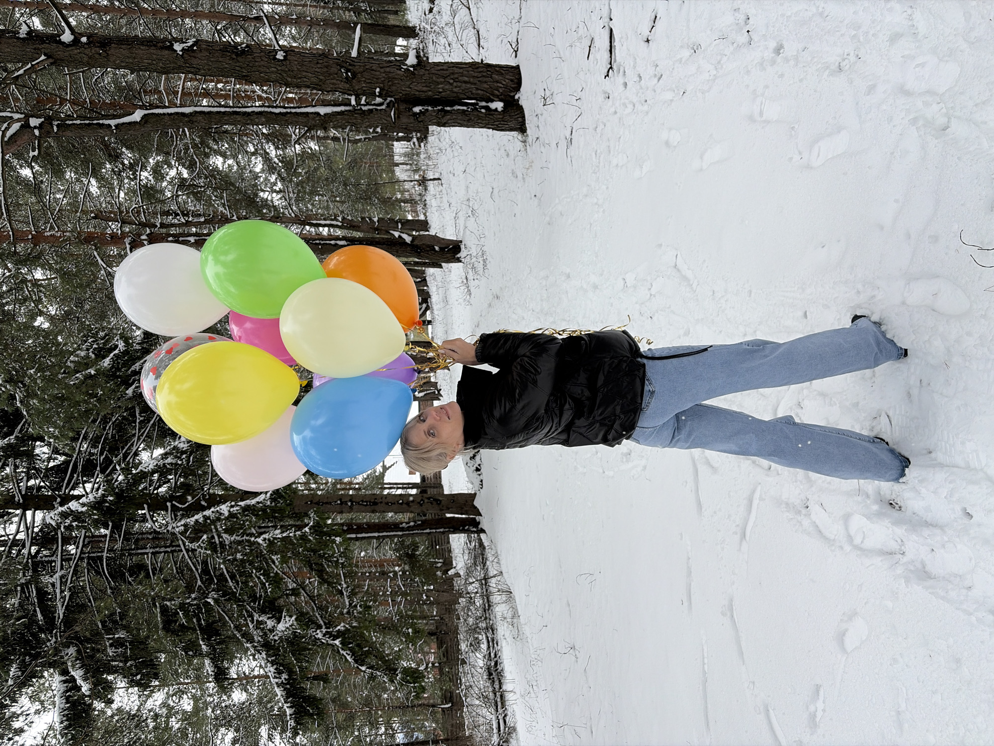 A woman stands in a snow-covered area with trees, holding a bunch of colorful balloons in Прикамский, Russia. She wears a black jacket and light blue jeans. The balloons are various colors including white, green, orange, yellow, pink, light blue, and a patterned clear balloon. The trees are visible in the background with snow on their branches.