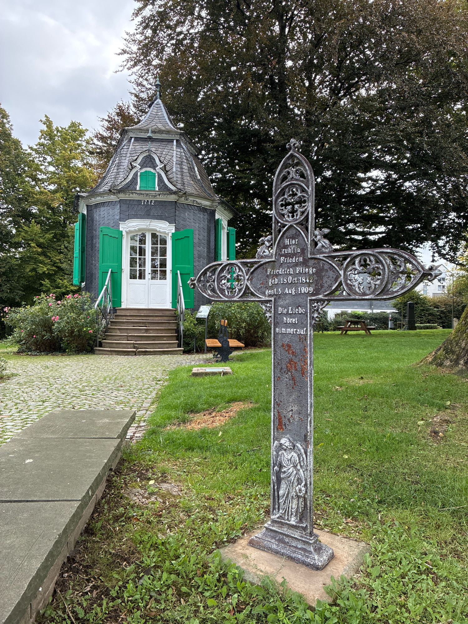 The image captures a gravestone in the form of a decorative cross, situated in a cemetery in Radevormwald, Germany. The cross is ornate, featuring intricate scrollwork and small angelic figures. Inscriptions on the cross are partially legible, indicating a person named Friedrich Fliege, born October 18, 1842, and died August 5, 1909. Below this, text reads "Die Liebe höret nimmer auf," meaning "Love never ceases."
In the background, a small, octagonal building with a distinctive grey, mansard-style roof and bright green shutters is visible. The building has white double doors and a set of stone steps leading up to it. The year "1772" is inscribed above the entrance. The surrounding environment is a well-maintained cemetery with lush green grass, mature trees, and cobblestone pathways. Other gravestones and a picnic table are present in the background. The sky is overcast, suggesting a cloudy day.