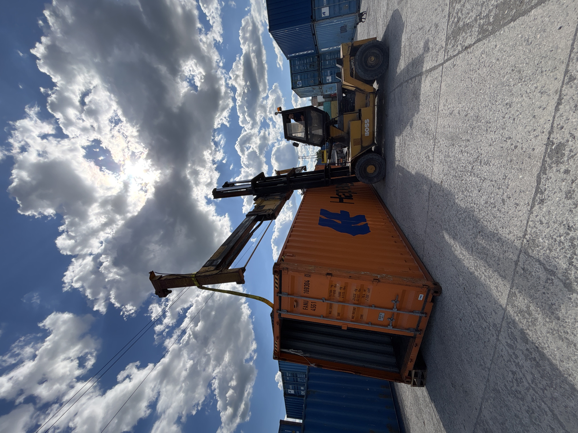 The image shows a yellow "BOSS" brand forklift lifting an orange shipping container at what appears to be a shipping yard. The container is marked with the code "FANU 169304 0 4561". Blue shipping containers are stacked in the background. The setting is an outdoor lot with a concrete surface. The sky is visible with a mix of blue and white clouds. The location in the city of Devnya, Bulgaria is not verifiable from the image.