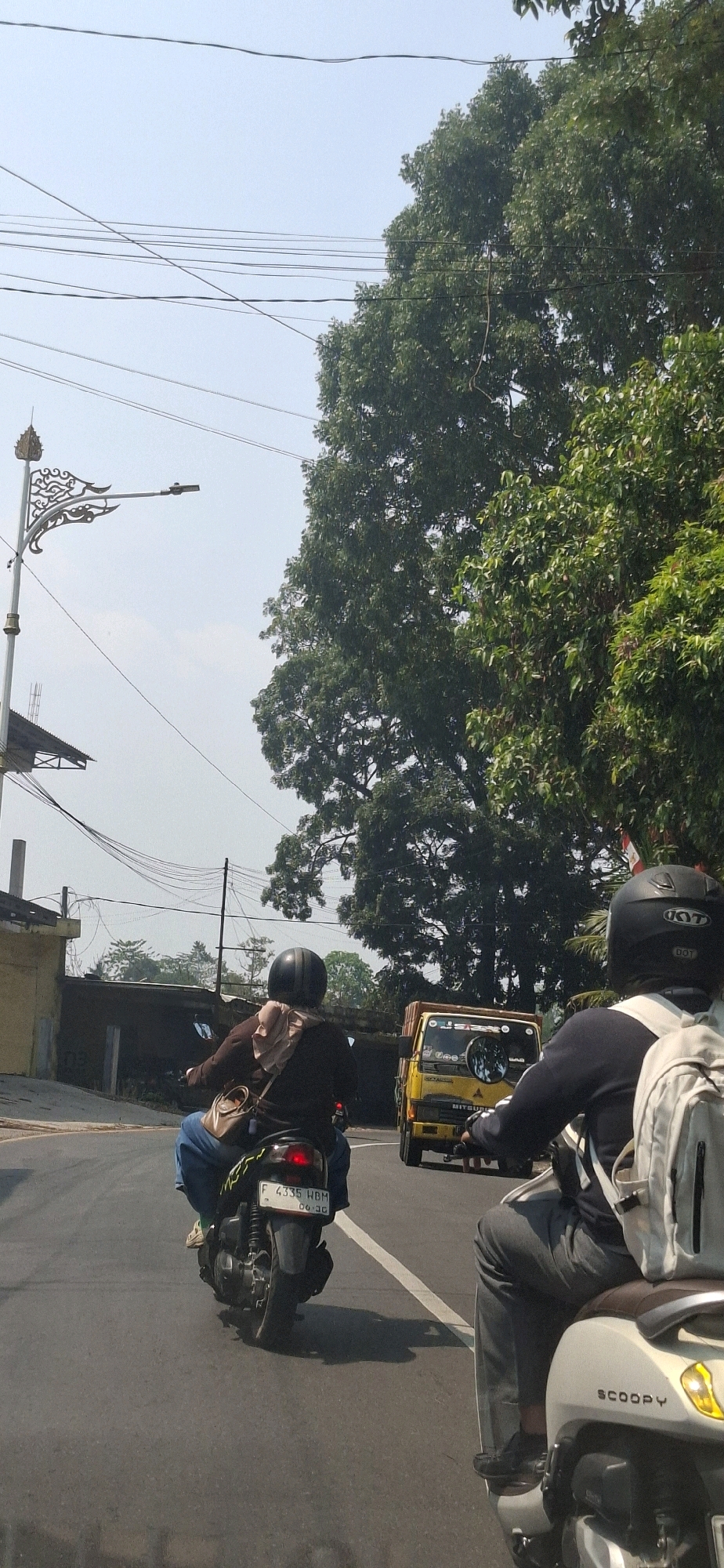 In Cilaku, Indonesia, two individuals are riding motorcycles on a paved road. The first motorcycle, a black scooter with a white license plate displaying "F 4335 HBM", is ridden by a person wearing a dark jacket and blue jeans, with a light-colored headscarf and a brown shoulder bag. The second motorcycle, a white scooter labeled "SCOOPY", is ridden by a person in a dark long-sleeved shirt, grey pants, and a black helmet with white accents. A yellow truck with a spare tire mounted on its rear is visible in the background, also on the road. The environment includes a large tree with dense green foliage to the right, a street lamp with an ornate metal design on a pole to the left, and power lines crisscrossing the hazy sky. Buildings with yellow and dark exteriors are present on the left side of the road. The lighting suggests a daytime scene with clear skies.