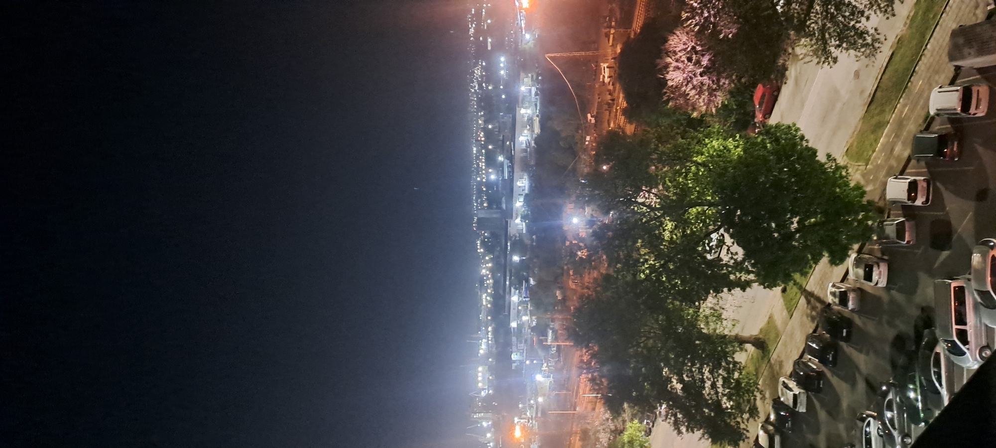 The image shows a nighttime view from an elevated position, possibly a high-rise building, overlooking the city of Burgas, Bulgaria. The left side of the image is dominated by a dark, featureless night sky. The city lights stretch across the horizon, creating a visual line of illumination. Prominent sources of light indicate industrial or port areas with brighter, more intense light clusters. A line of trees and a parking area filled with cars are visible in the foreground on the right side.