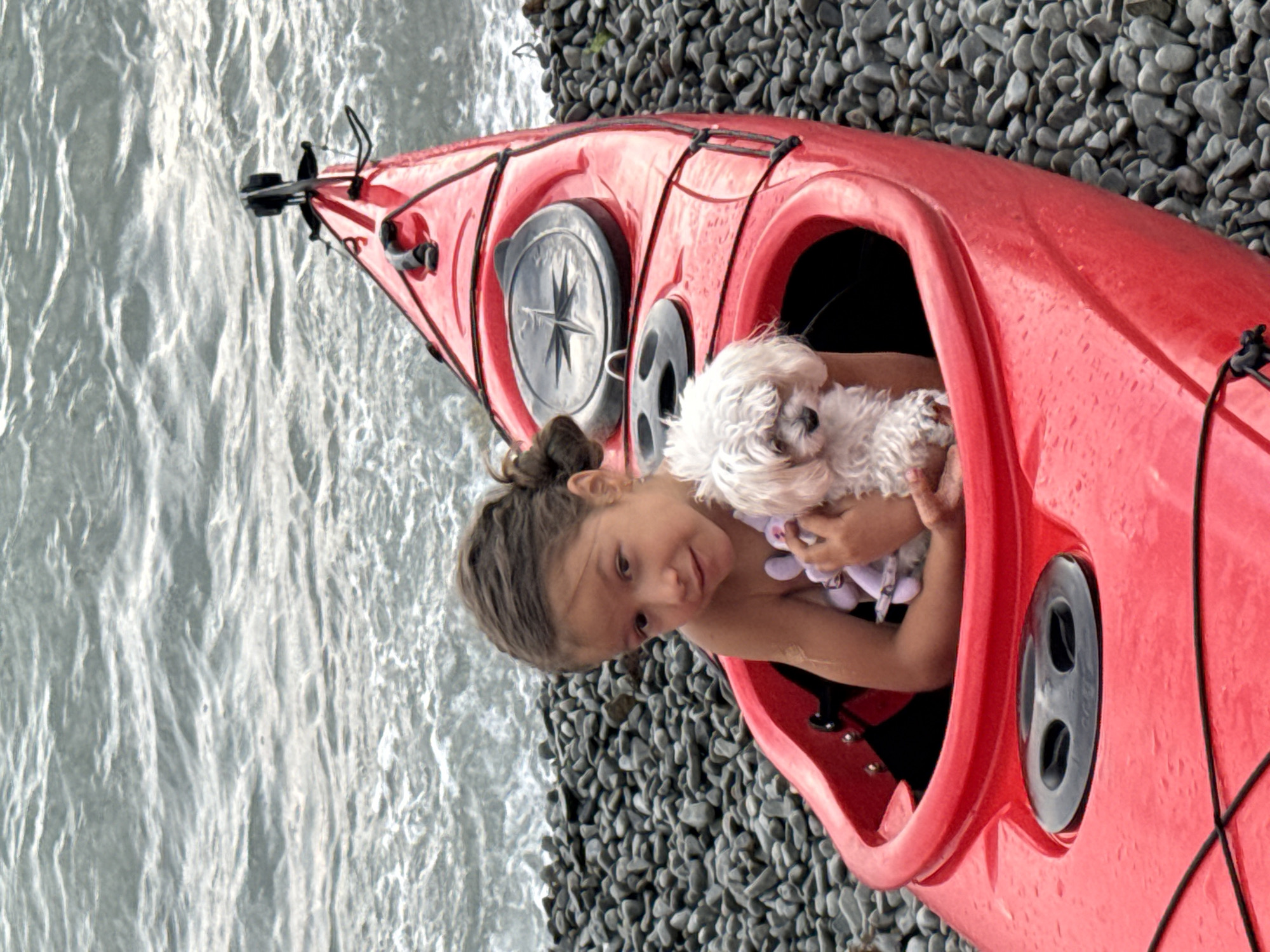 In Partenit, Ukraine, a young girl sits in a red kayak on a rocky shore. She holds a small, white dog. The water is choppy and grey. The girl is looking at the camera and smiling. The dog is looking down. The kayak is filled with the girl and the dog. There is a compass on the kayak. The scene is bright and sunny.