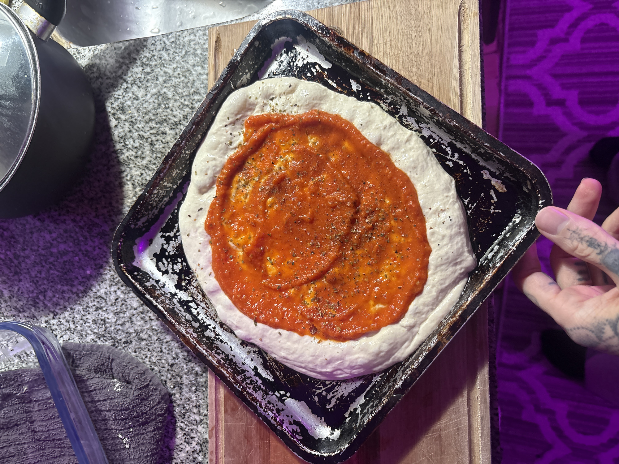 A pizza in a square baking pan sits on a wooden cutting board. The pizza dough is topped with a red tomato sauce and some dried herbs. A person's hand is visible in the right side of the image. A pot is on the left and a grey towel. The background contains marble countertops, and a purple ornate pattern. The location is unknown and cannot be determined.