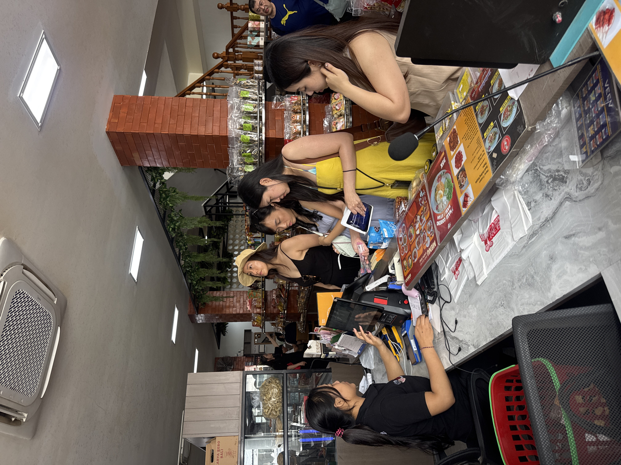 In Kuta, Indonesia, this indoor scene depicts a bustling checkout counter in what appears to be a souvenir or food shop. Four women and one man are visible. A female cashier, wearing a black uniform with a logo on the left sleeve, is seated and interacting with a computer terminal. Her right hand is on the keyboard, and her left hand is near a cash register.
Three women are standing at the counter. One, wearing a straw hat and a black tank top, is looking upwards. The woman next to her, in a yellow sundress, is holding a tablet or e-reader and showing it to the woman on her left, who is wearing a light blue dress. This woman is holding cash in her hand. Another woman, wearing a beige tank top, stands to the right of the cashier, facing the counter and appearing to be listening.
Behind the counter, shelves are stocked with various packaged goods, including snacks and souvenirs. A brick pillar is visible to the right, supporting the ceiling where an air conditioning unit and fluorescent lights are mounted. A wooden staircase with a railing is also present in the background. A man in a blue t-shirt with a yellow logo is standing near the stairs, looking towards the checkout area. On the counter, menus are displayed, and shopping bags are neatly stacked. The counter surface has a marble-like pattern, and various electronic devices and office supplies are scattered across it.