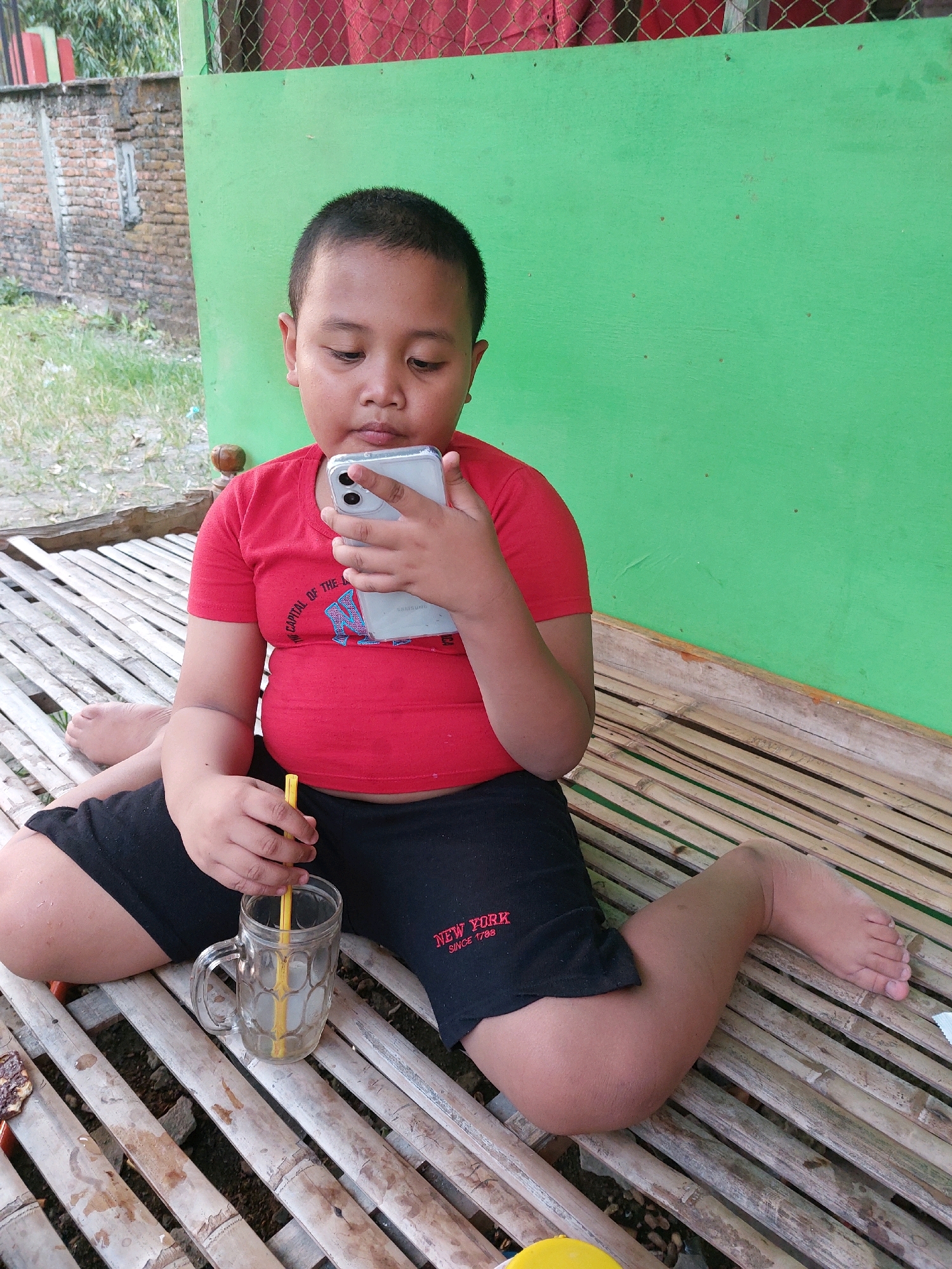 A young boy, likely Indonesian, is seated on a bamboo matting outdoors. He is wearing a red t-shirt and black shorts. The boy is holding a silver smartphone in his hands and looking at the screen. Next to him, on the bamboo mat, is a clear glass mug with a yellow straw and a light-colored liquid. The background features a green wall and a red mesh fence. The scene appears to be in a casual, outdoor setting, possibly a yard or patio, in Canga'an, Indonesia.