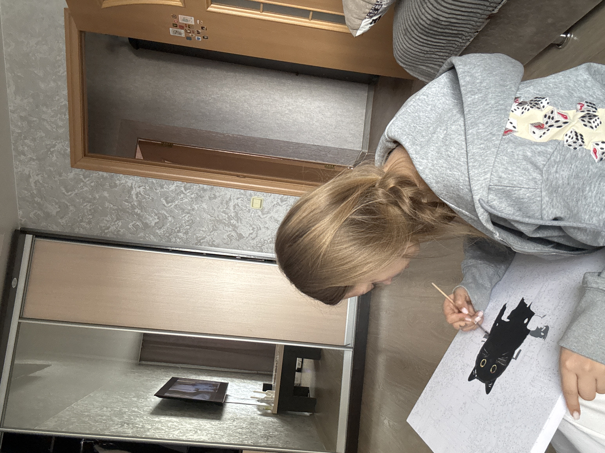 An indoor, vertically oriented image captures a young woman engrossed in painting a paint-by-numbers kit in a domestic setting, identified as Chaikovsky, Russia. The woman, with light-colored hair, is seen from a high angle, her gaze fixed on the canvas. She wears a grey hooded sweatshirt featuring a pattern of dice and holds a slender paintbrush, applying black paint to outline a black cat with prominent yellow eyes on the white canvas.
The background reveals a light wooden door frame or mirror frame adorned with small, unreadable magnets or stickers. The room's walls are covered in a light grey, subtly patterned wallpaper. To the left, a modern sliding wardrobe with light wooden panels and reflective surfaces is prominent. A small, dark rectangular device, possibly a phone or tablet, lies on the wardrobe's reflective top. Further into the background, a grey striped pillow with the visible text "300" suggests a bed or seating area. The scene portrays a calm, focused activity during what appears to be daytime within a well-kept home environment.