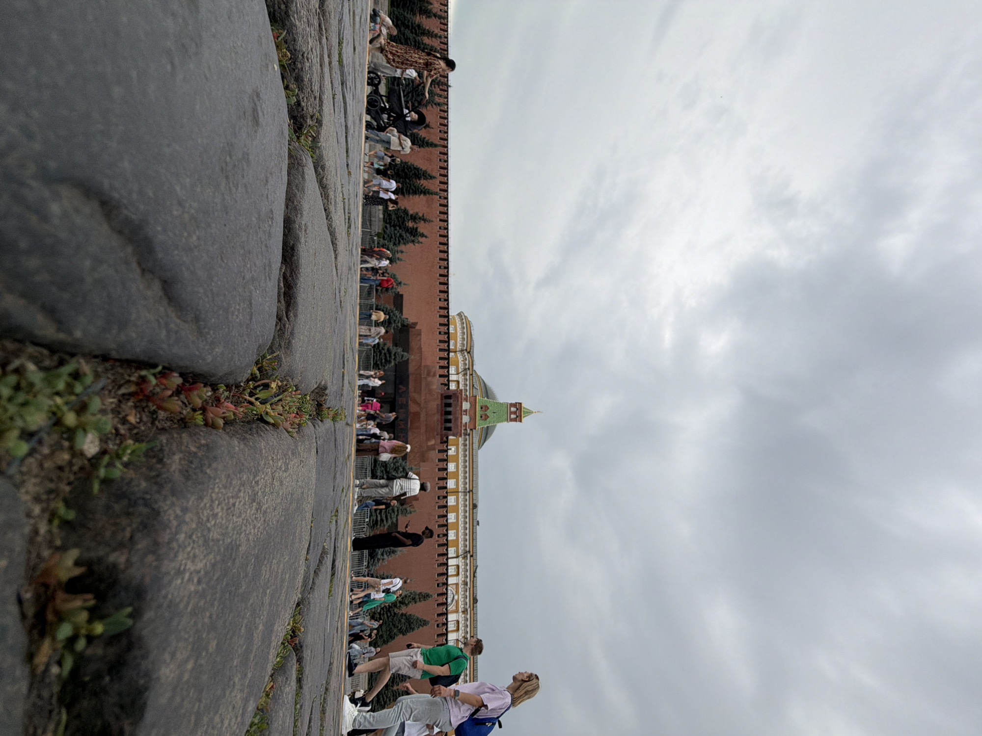 The image depicts a ground-level view of a cobblestone path with several people walking along it. In the background, a large, red brick building with a green-domed tower is visible. The building appears to be a historical landmark, possibly part of a fortress or palace complex. Pine trees are planted in rows along the base of the building. The sky is overcast, suggesting a cloudy or possibly rainy day. The people are dressed in casual summer clothing, and some are carrying bags, indicating they are likely tourists or visitors. The overall atmosphere seems relaxed and casual, with individuals and small groups moving in the same direction. The low angle of the shot emphasizes the texture of the cobblestones in the foreground.