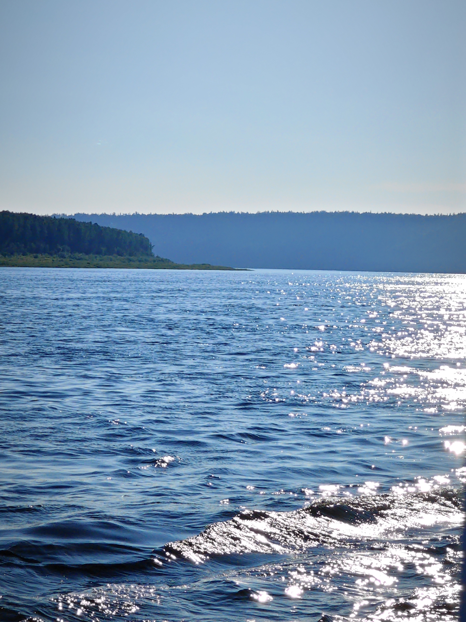 This is a medium-shot, naturalistic photograph of a large body of water, likely a river or lake, with a distant tree-lined shore and a clear sky. The water is a deep blue and shows small ripples and waves, reflecting the sunlight in a shimmering pattern across the surface. The shore in the distance is covered in dense green trees and appears as a dark, undulating silhouette against the lighter blue sky. The overall scene suggests a calm, natural environment, possibly captured during daytime on a sunny day. The image does not contain any people or man-made objects, and therefore, cannot be linked to a specific location within the city of Bor, Russia.