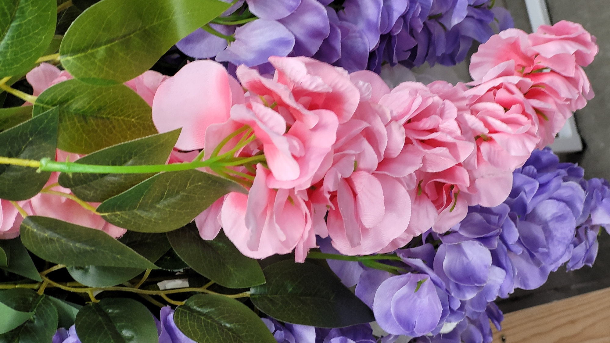 The image shows a close-up of artificial flowers. There are cascading blooms in pink and purple, interspersed with green leaves. The environment is indistinct. No people, subjects, objects, actions, or interactions are readily discernable other than what is described. The location is unknown.