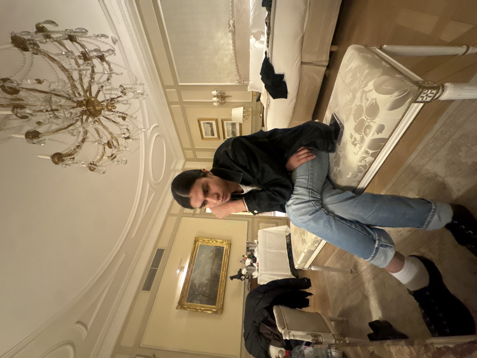 A young individual with dark, long hair pulled back is seated on an ornate, upholstered bench with a floral pattern and gilded accents. They wear a white t-shirt, a dark denim jacket, light blue jeans cuffed at the ankles, white socks, and dark shoes. Their right hand supports their chin, and their left hand rests on their lap, next to a dark-colored smartphone placed on the bench.
The scene is an interior room, characteristic of a luxury hotel suite, located in Paris, France. The room features a white ceiling with classical decorative moldings and a large, multi-tiered brass and crystal chandelier. The walls are light-colored with paneling. A large bed with a light-colored headboard and white bedding is visible on the right, with dark clothing draped across it. To the left, a small table covered with a white tablecloth holds a champagne bucket containing two bottles, a glass, and other items. A dark jacket is draped over an adjacent chair. A large landscape painting in a gold frame hangs on the left wall, above a dark decorative horse figurine. Two smaller framed artworks are stacked on the right wall. The flooring consists of light-colored wood with a patterned rug. An air conditioning vent is present on the upper left wall.