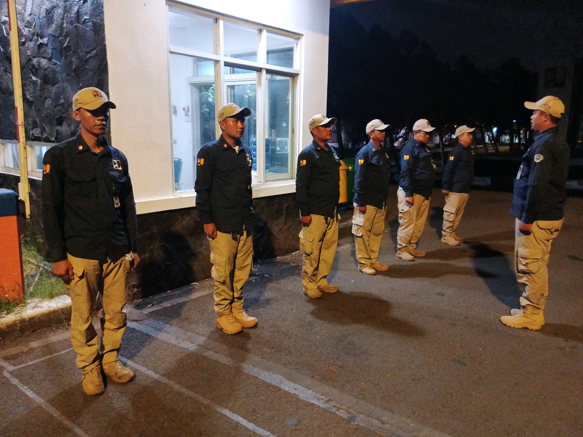 Six men in matching uniforms and caps are standing in a line outdoors at night. The men are wearing dark long-sleeved shirts with orange patches on the sleeves, tan cargo pants, and tan combat boots. They are standing on asphalt pavement, with white lines painted on it. Behind them is a building with large windows and a dark exterior wall. The setting appears to be a secure compound or institution. The overall scene suggests a security or patrol detail on duty.