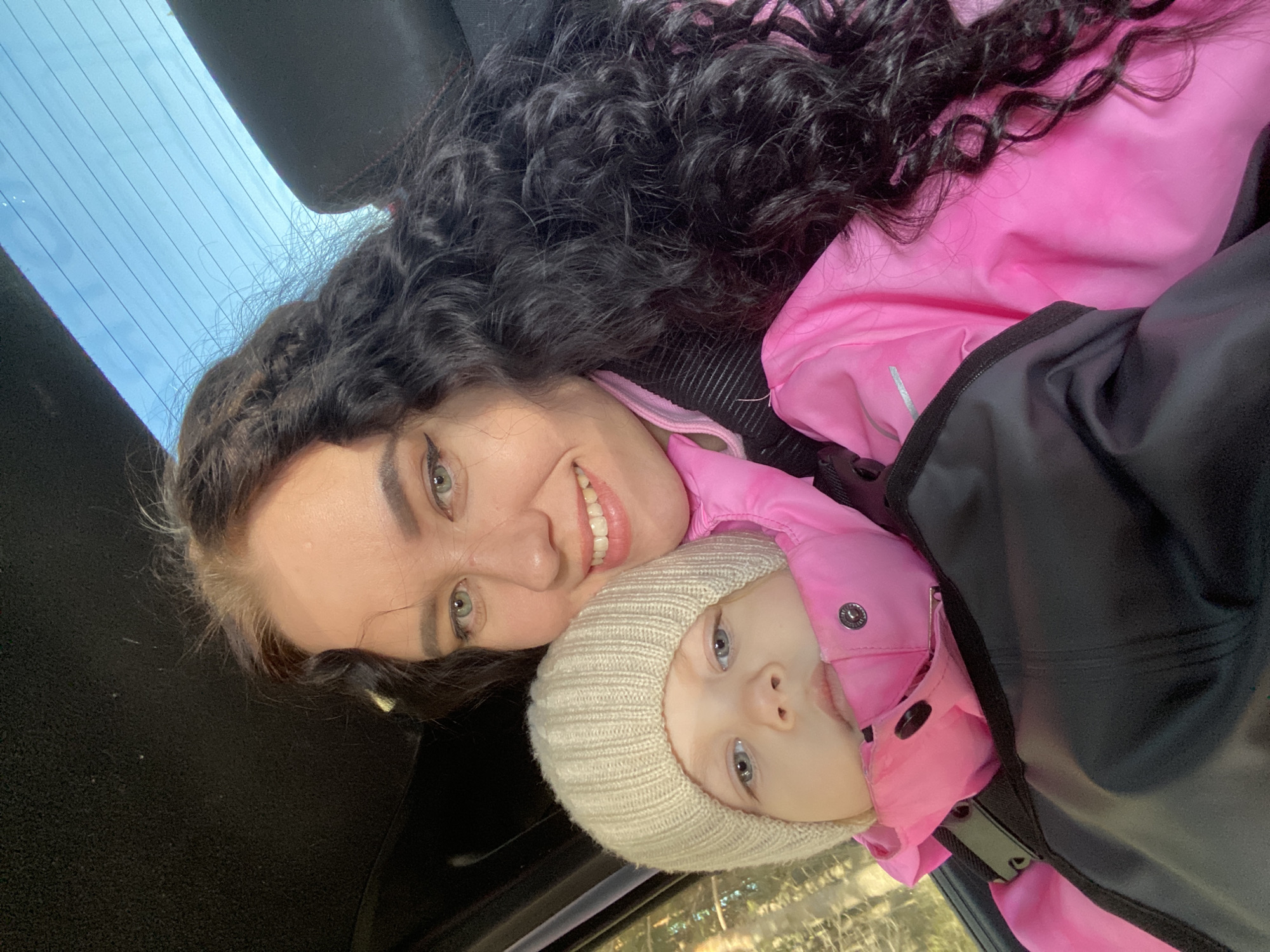 A photograph depicts a woman and a young child in an indoor setting, likely a vehicle, based on the visible car interior elements. The woman has dark, curly hair and is wearing a pink top. She is smiling and looking directly at the camera. The child, positioned close to the woman, has light hair and is wearing a knitted hat and a pink jacket. The background includes a partially visible car window. While the image does not provide explicit clues to confirm the exact location as Chaikovsky, Russia, this cannot be ruled out completely.
