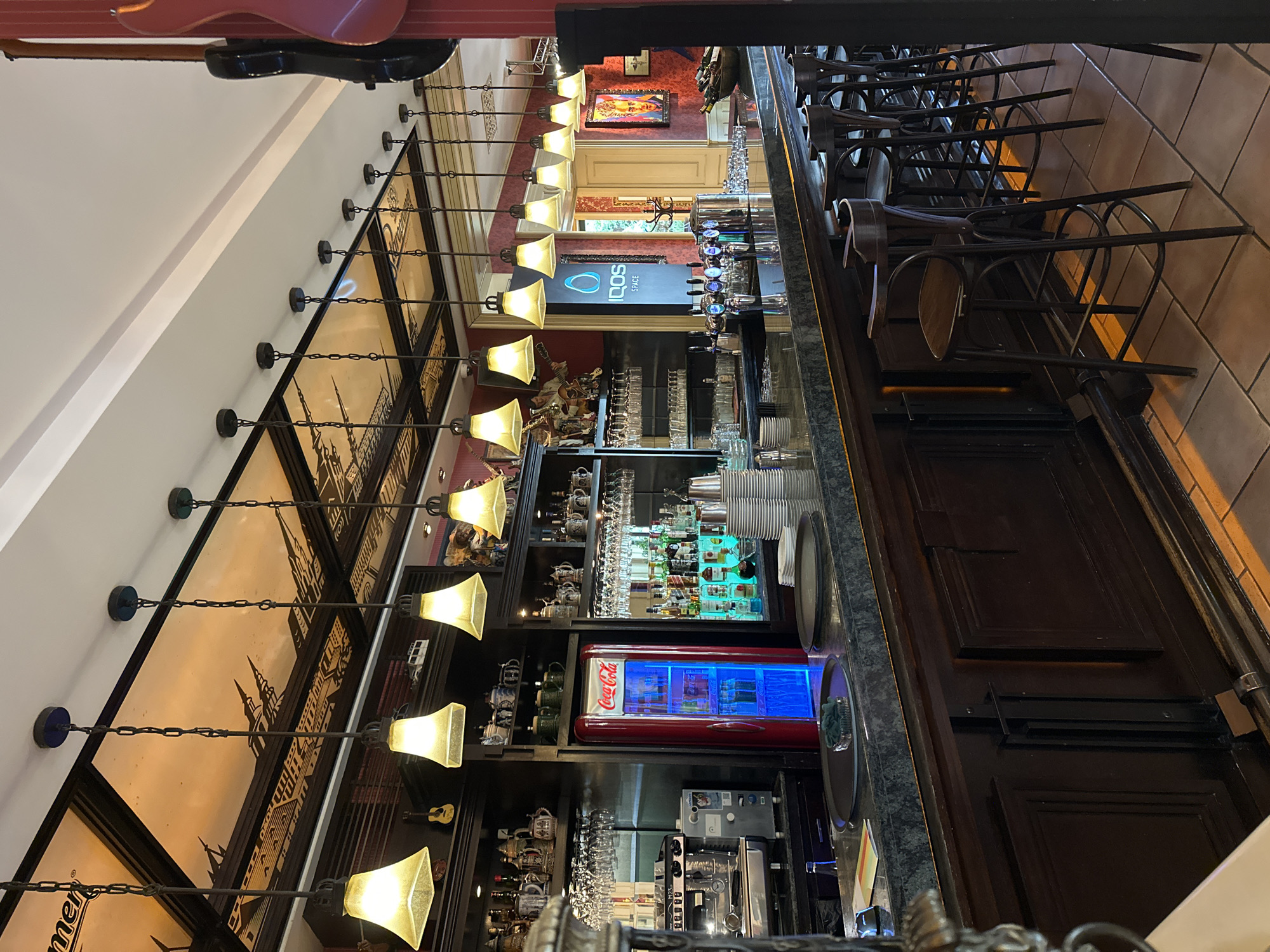 The image depicts the interior of a bar, characterized by a long, polished countertop and a row of high stools. Above the bar, numerous pendant lights with amber-colored glass shades illuminate the space, hanging from a chain-linked system. The ceiling features a decorative panel with a cityscape motif. Behind the bar, shelves are stocked with various bottles, glassware, and decorative items, including ceramic mugs and figurines. A bright red Coca-Cola refrigerator is prominently displayed. To the right, a black acoustic guitar is mounted on the wall. The bar area appears clean and organized, with stacks of plates and glasses ready for service. The overall atmosphere suggests a lively and well-equipped establishment. The presence of the Coca-Cola refrigerator and the general ambiance suggests this could be a bar or restaurant. The specific location in Subotica, Serbia cannot be determined from the image alone.