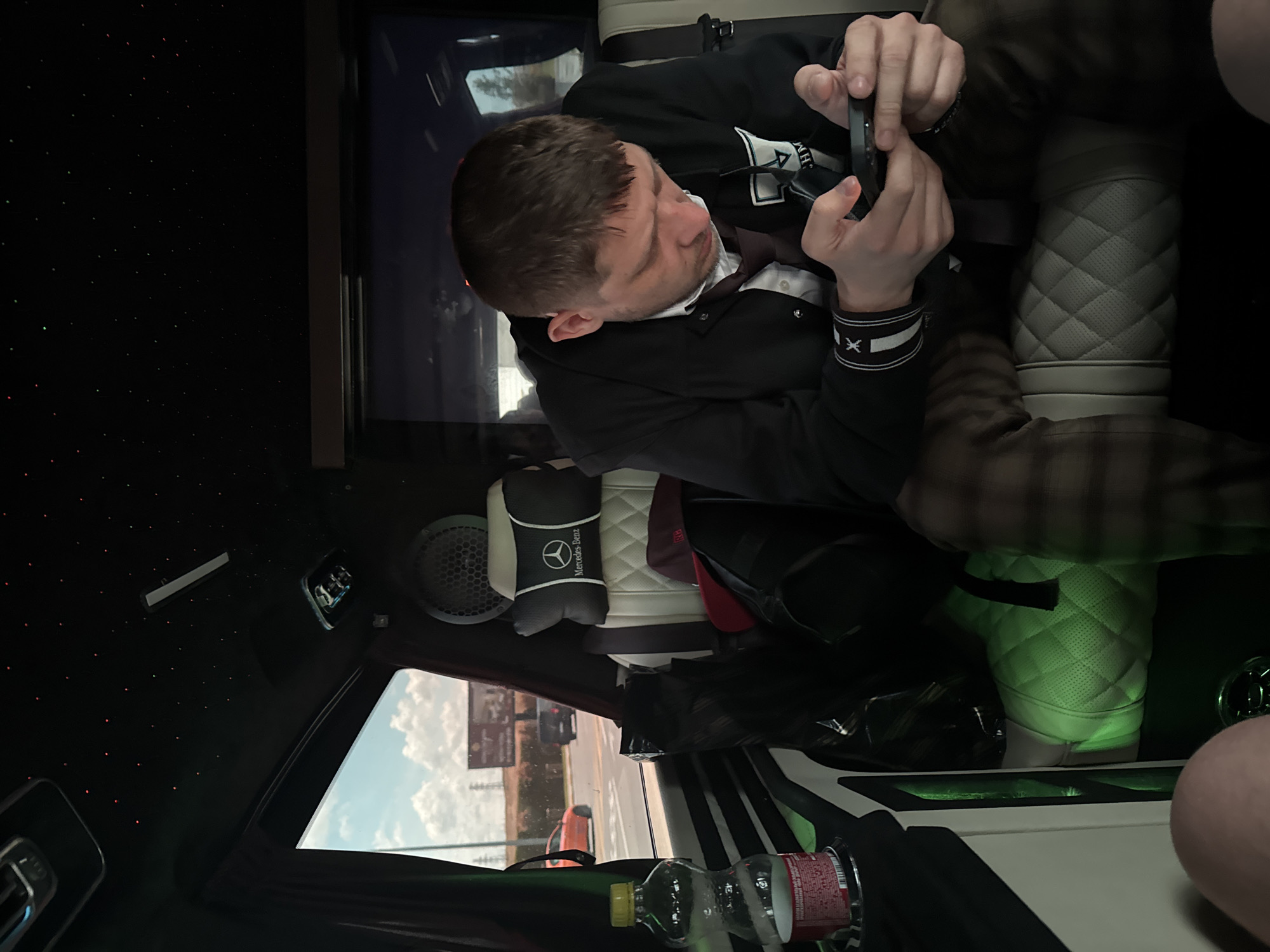 The image shows a man sitting inside a luxury vehicle, likely a Mercedes-Benz van, as indicated by the headrest. He is wearing a dark jacket over a white shirt and a tie, and plaid pants. The man is looking down at his phone, which he is holding in his hands. The interior of the vehicle features a quilted white seat and green ambient lighting. The ceiling is black with small red lights, resembling a starry sky. Through the window, a street scene is visible with cars and a billboard, suggesting an urban or semi-urban environment. The location is identified as Arnavutköy, Turkey.