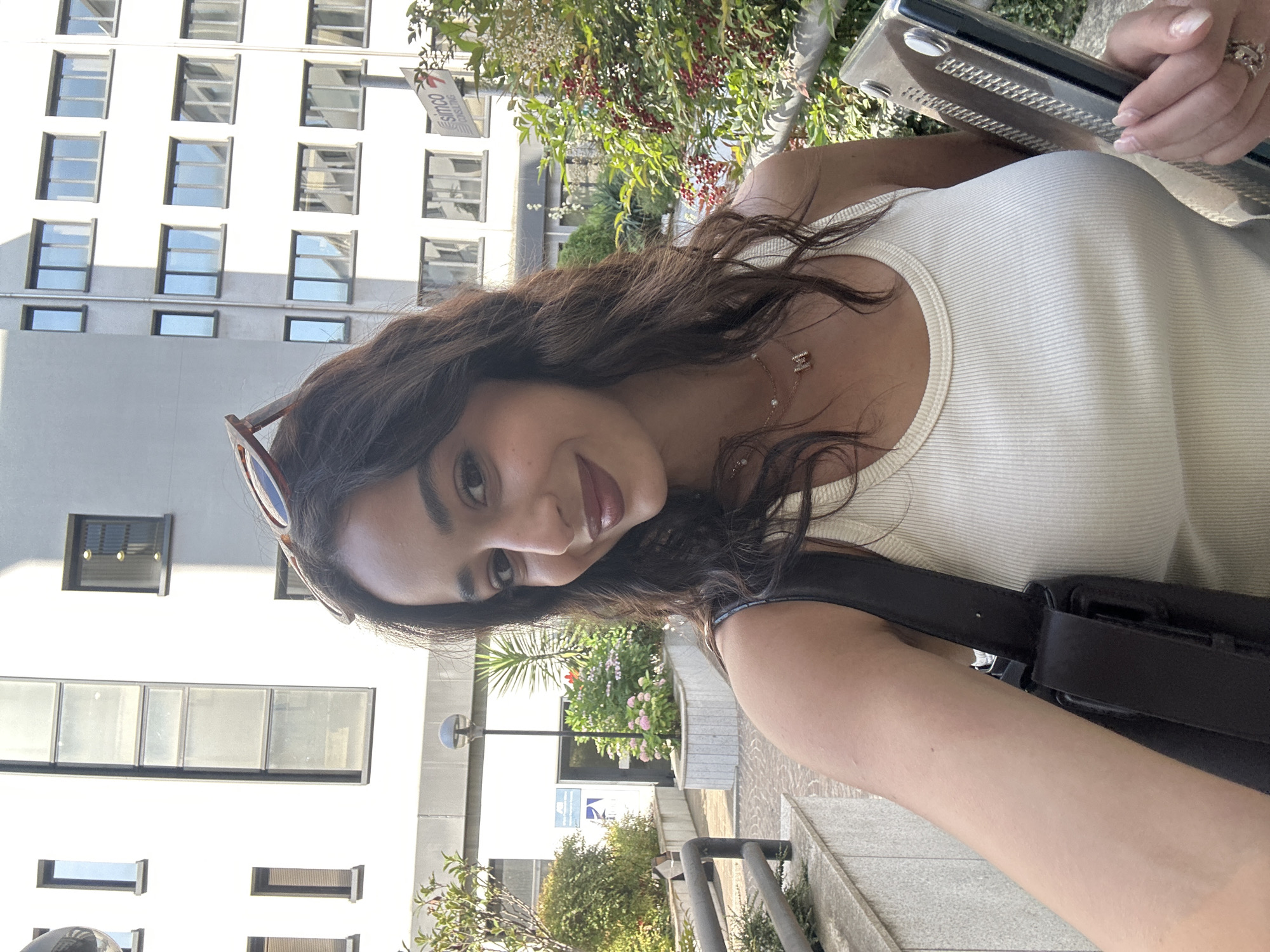 A woman with long, dark curly hair, wearing a white tank top and sunglasses atop her head, is holding a laptop in a clear plastic case. She is standing outdoors in front of a modern building with many windows. There are trees and bushes nearby. The location is Milan, Italy.