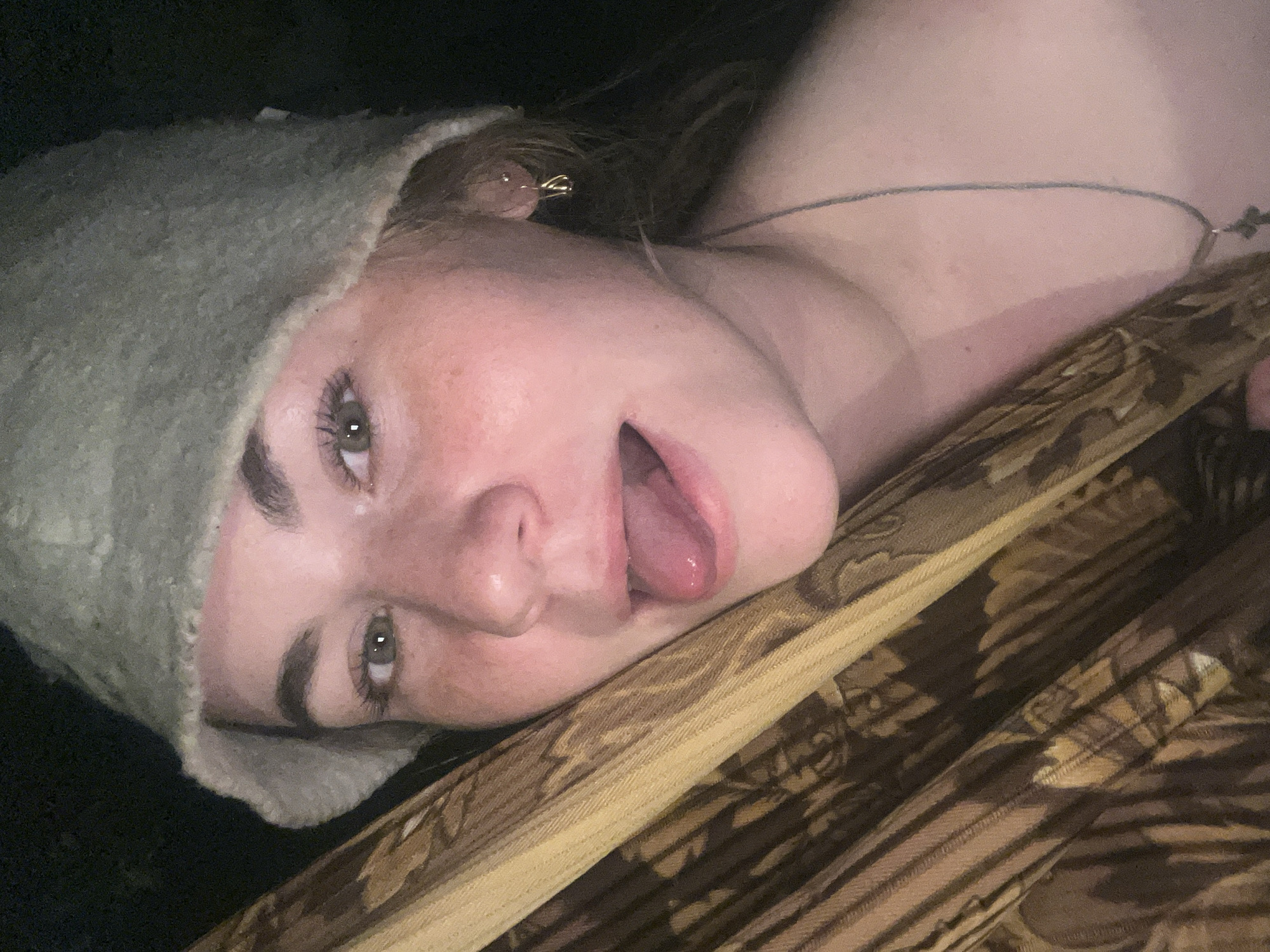 A close-up, tilted image shows a young woman lying down. She is wearing a soft, possibly wool, hat. Her mouth is slightly open, and her tongue is visible. A thin necklace is around her neck. She appears to be resting on or against a patterned fabric. The location is Сосново, Russia, but the image does not contain any details that confirm the location.