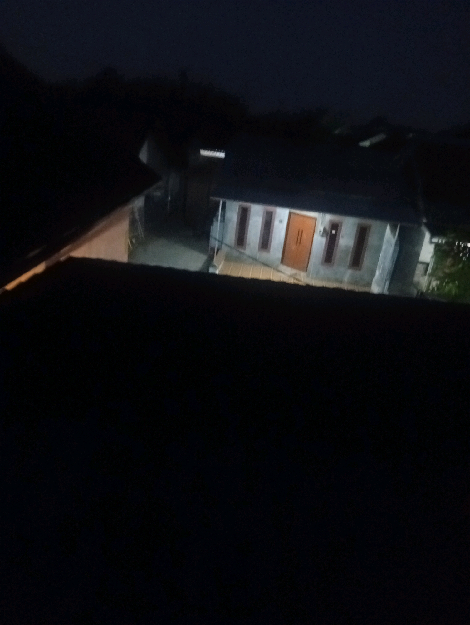 This is a dark, low-light image of a house at night. The house appears to be a single-story dwelling with a gray facade. It has a prominent wooden double door, painted orange or brown, and several rectangular windows. The windows are tall and narrow, with dark frames. The house is illuminated by interior lights, which cast a warm glow from the windows and through the doorway. The foreground of the image is dominated by a dark, silhouetted roof structure, suggesting the photograph was taken from a higher vantage point, possibly another roof or elevated area. The background shows the dark outlines of trees and possibly other structures, indicating a residential area. The overall impression is one of a quiet, dimly lit neighborhood at night.