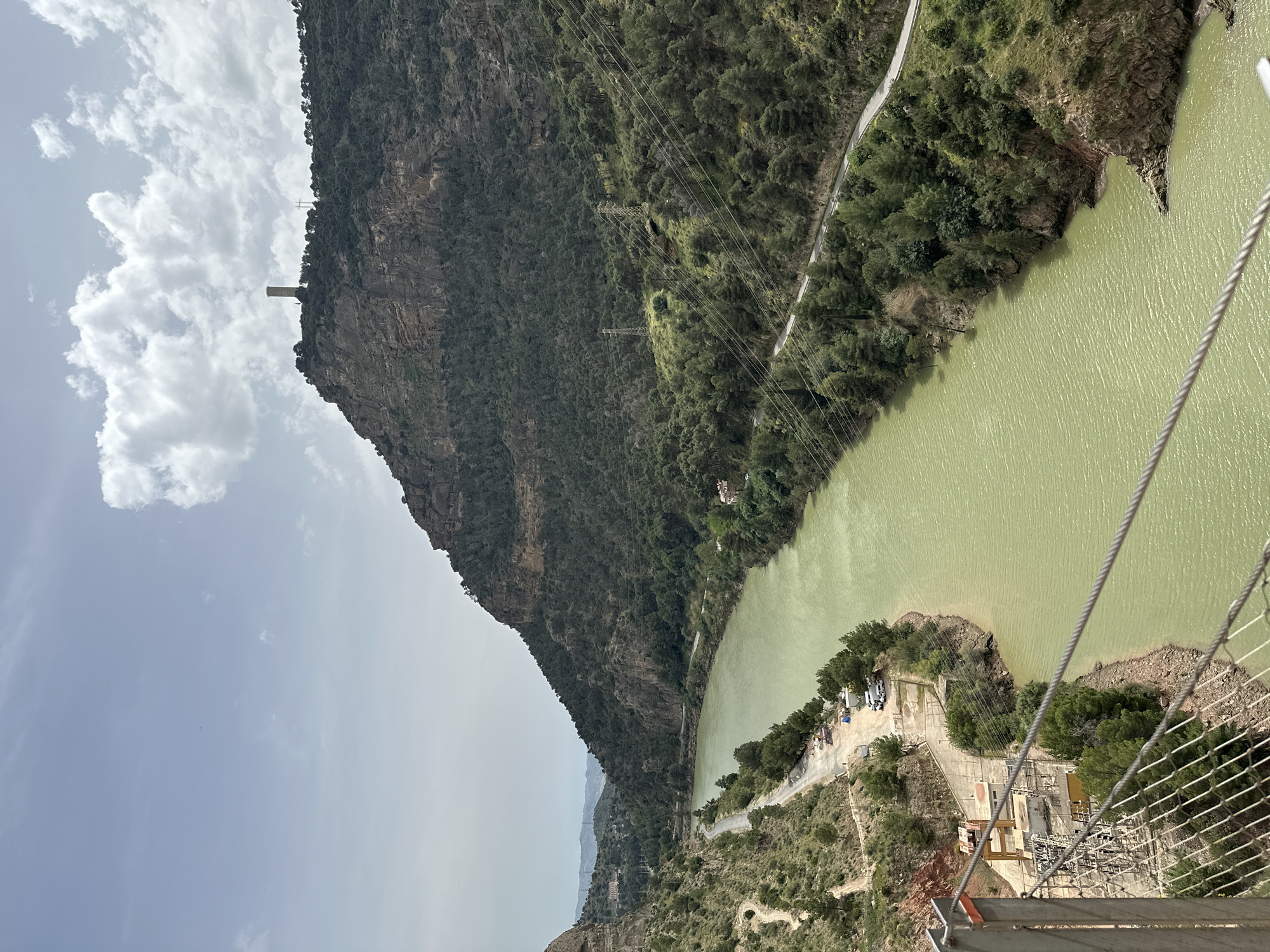 The image presents a dramatic aerial view of a mountainous valley with a winding body of greenish-yellow water, likely a reservoir or river. The photograph is taken from a high vantage point, possibly a suspension bridge or a cliffside path, evidenced by the metal cables and mesh fence visible in the extreme right foreground.
On the left, a towering, rugged mountain face rises sharply, densely covered in dark green foliage and rock formations. A distinctive, slender stone tower stands prominently atop this cliff, silhouetted against a cloudy sky. The sky is partially covered with white, scattered clouds.
The right side of the frame features the water body's shore, with a winding road tracing its edge through sparse vegetation. Below the viewer, at the water's edge, a complex of buildings and industrial infrastructure, possibly a power station or a water management facility, is visible. Multiple power lines span across the valley, supported by pylons embedded in the forested slopes. The scene depicts a grand natural landscape integrated with significant human engineering, characteristic of the area near Álora, Spain. There are no people or visible text. The time of day appears to be daytime, under partly cloudy skies.