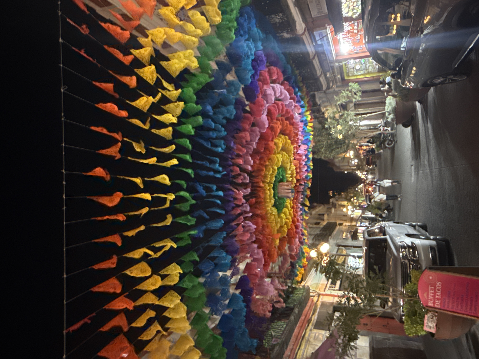 This image captures a vibrant street scene at night, likely in Puebla, Mexico, dominated by an elaborate overhead display of colorful pennants or flags. These decorations, reminiscent of "papel picado," are strung across the width of the street, forming a long, arched canopy arranged in a rainbow spectrum of red, orange, yellow, green, blue, purple, and pink hues.
The street below is bustling with activity and illuminated by numerous warm-toned streetlights and lights from the surrounding buildings. The architecture features multi-story urban structures, some with balconies, indicative of an older city center. Several vehicles are visible, including a dark car in the mid-ground on the right and a lighter-colored SUV further down the street, alongside a parked motorcycle.
Several indistinct figures of people can be seen in the distance, walking or standing, suggesting a lively pedestrian area. Potted plants and small trees are placed along the sidewalks, adding greenery to the urban environment.
A prominent red sign in the foreground on the right clearly advertises "BUFFET DE TACOS" in large white letters. Below this, a list of various taco fillings is partially visible, including "PASTOR," "ARABE," "SUADERO," "CECINA," "CHULETA," "CARNITAS," "LONGANIZA," and "CAMPECHANO."
The time of day is night, with artificial lighting providing the main illumination. The weather appears clear. The overall atmosphere conveyed by the bright colors and active street scene is cheerful, festive, and inviting, suggesting an evening of leisure and dining.