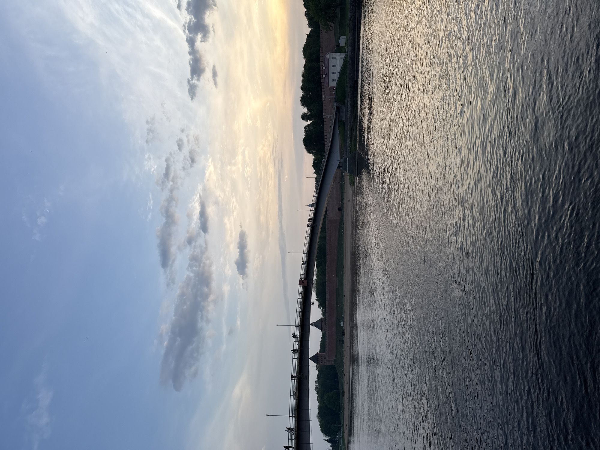 The image presents a long horizontal view of a river with reflections, bordered by structures. A bridge stands prominently in the scene. Buildings and trees are visible along the riverbank. The sky shows a combination of clouds and clear sections, indicating daytime.
Location: Veliky Novgorod, Russia. The image shows the Volkhov River with the pedestrian bridge leading towards the Novgorod Kremlin.