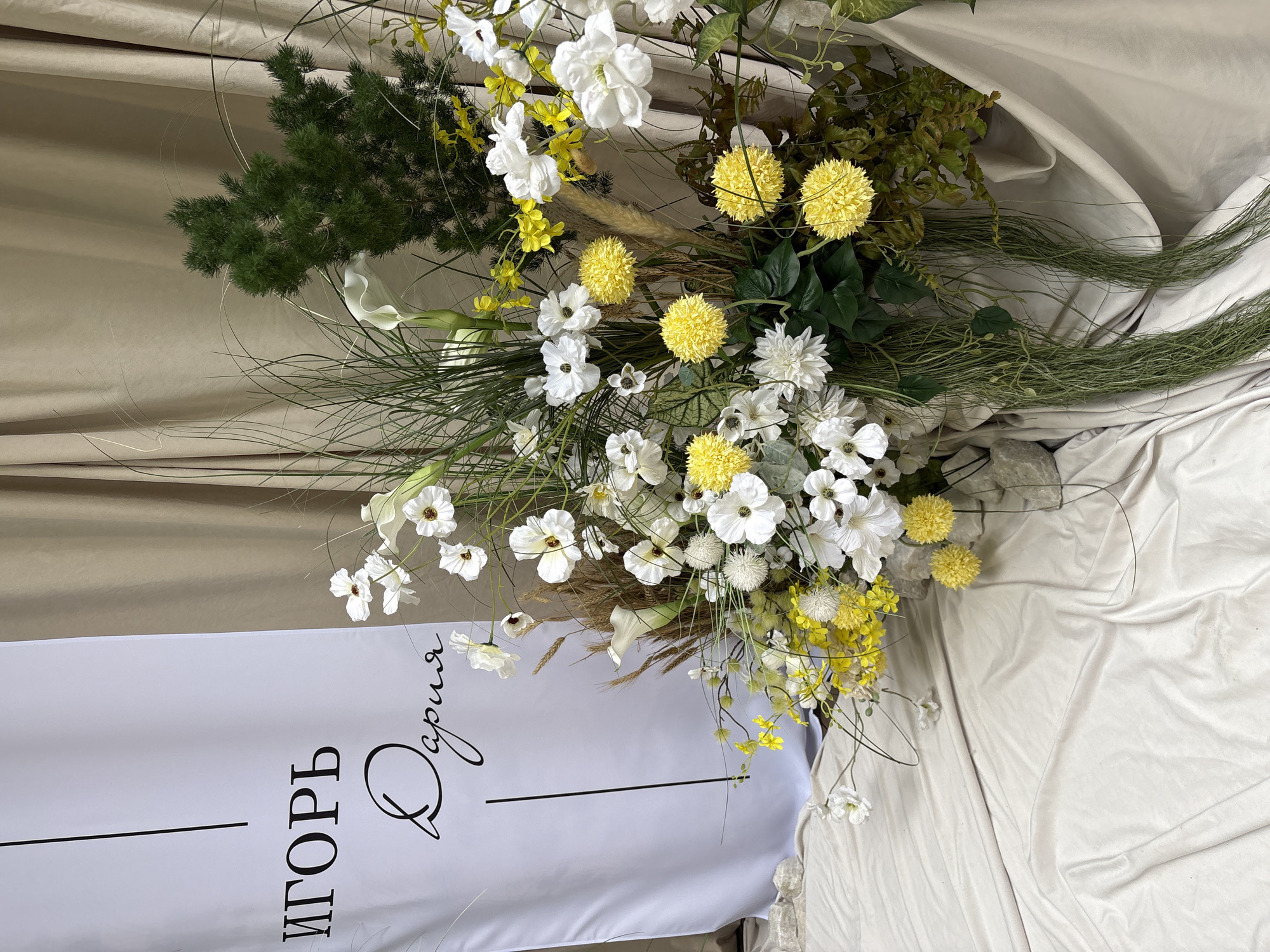 This is an indoor scene depicting a large floral display and a personalized banner, located in Perm, Russia. The central subject is a substantial floral arrangement composed primarily of white and yellow flowers with extensive green foliage. White flowers include several calla lilies and numerous multi-petaled blossoms, some with visible dark centers. Yellow flowers consist of spherical, ball-shaped varieties and clusters of small, delicate blooms on thin stems. The green elements feature tall, wispy grasses, dense bushy foliage resembling an evergreen, broad darker leaves at the base, and long, trailing tendrils. Also incorporated are several brown, feathery plumes. The arrangement is situated at the bottom-center and extends upwards and to the right. To the left of the floral display, a white fabric banner hangs vertically. It displays the name "ИГОРЬ" (IGOR) in black uppercase letters at the top, and "Дария" (Daria) in black script lettering below it, separated by a thin vertical black line. The background on the right and top consists of uniformly draped beige or light brown fabric curtains. The foreground and base of the display are covered with lightly wrinkled, draped light cream-colored fabric, upon which several small, irregular light-colored rocks are placed. No individuals or specific actions are depicted.