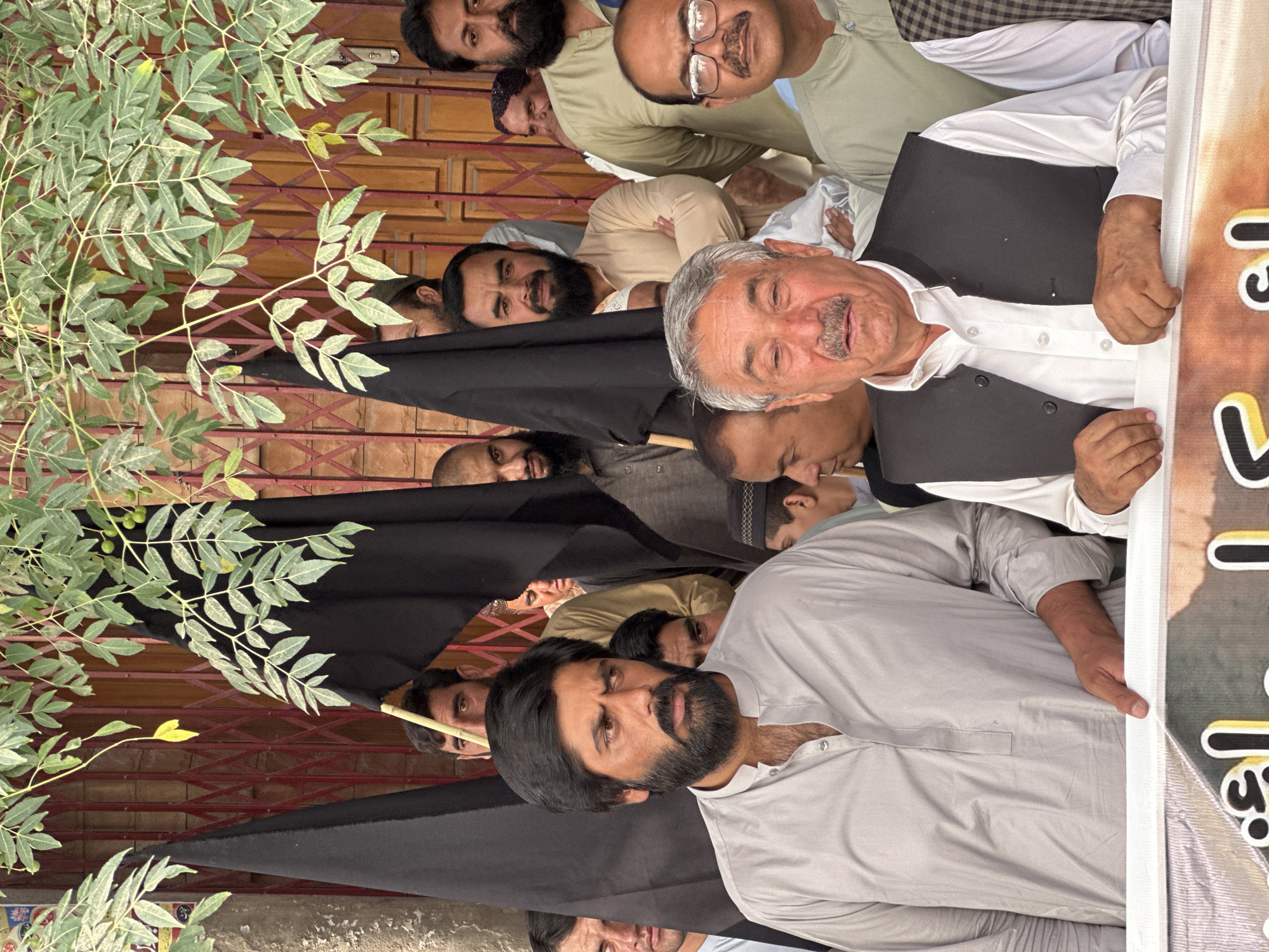 In Quetta City Tehsil, Pakistan, a group of men are gathered outdoors. The men are standing in front of a red metal gate and a building with wooden doors. Several men are holding black flags. Some of the men are looking towards the camera, while others are looking away. The image appears to be taken during the daytime. The overall mood of the image is serious. The men are dressed in traditional Pakistani clothing. A banner with text is visible at the bottom of the image.