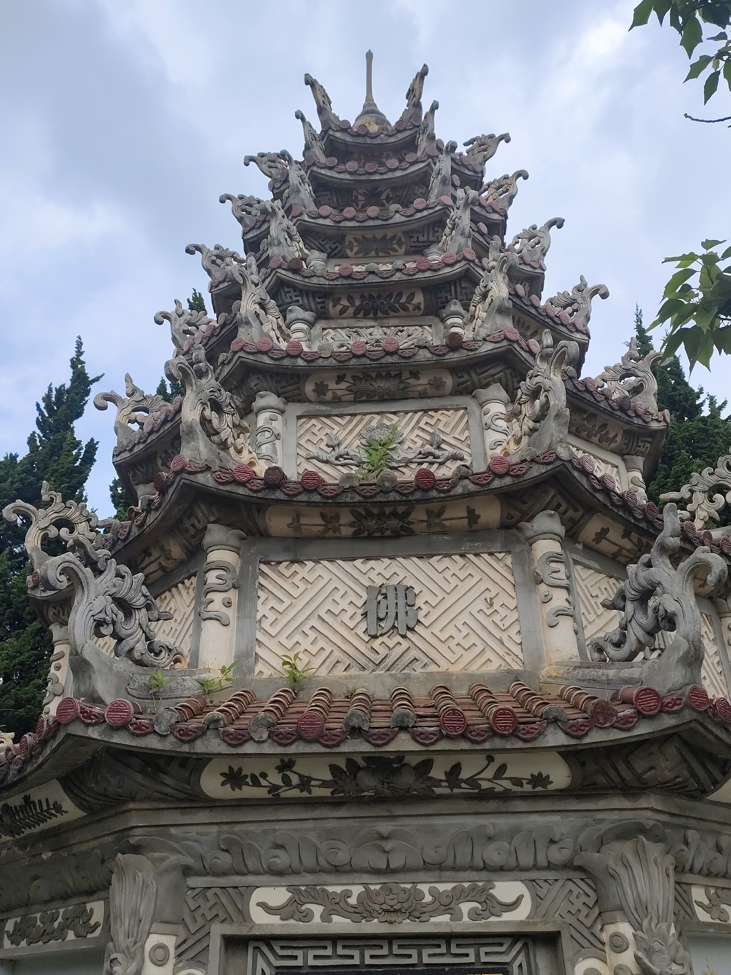 The image shows the upper portion of a multi-tiered, ornate pagoda-like structure. The structure is primarily light gray and white, with red-tiled roofing accents. It is heavily decorated with carved details, including dragon-like figures, floral motifs, and geometric patterns. The pagoda has several levels, each smaller than the one below, creating a tiered effect. In the background, there are tall, dark green trees and a cloudy sky. There is a Chinese character in the center. The location is Cam Ly - Đà Lạt, Vietnam.
