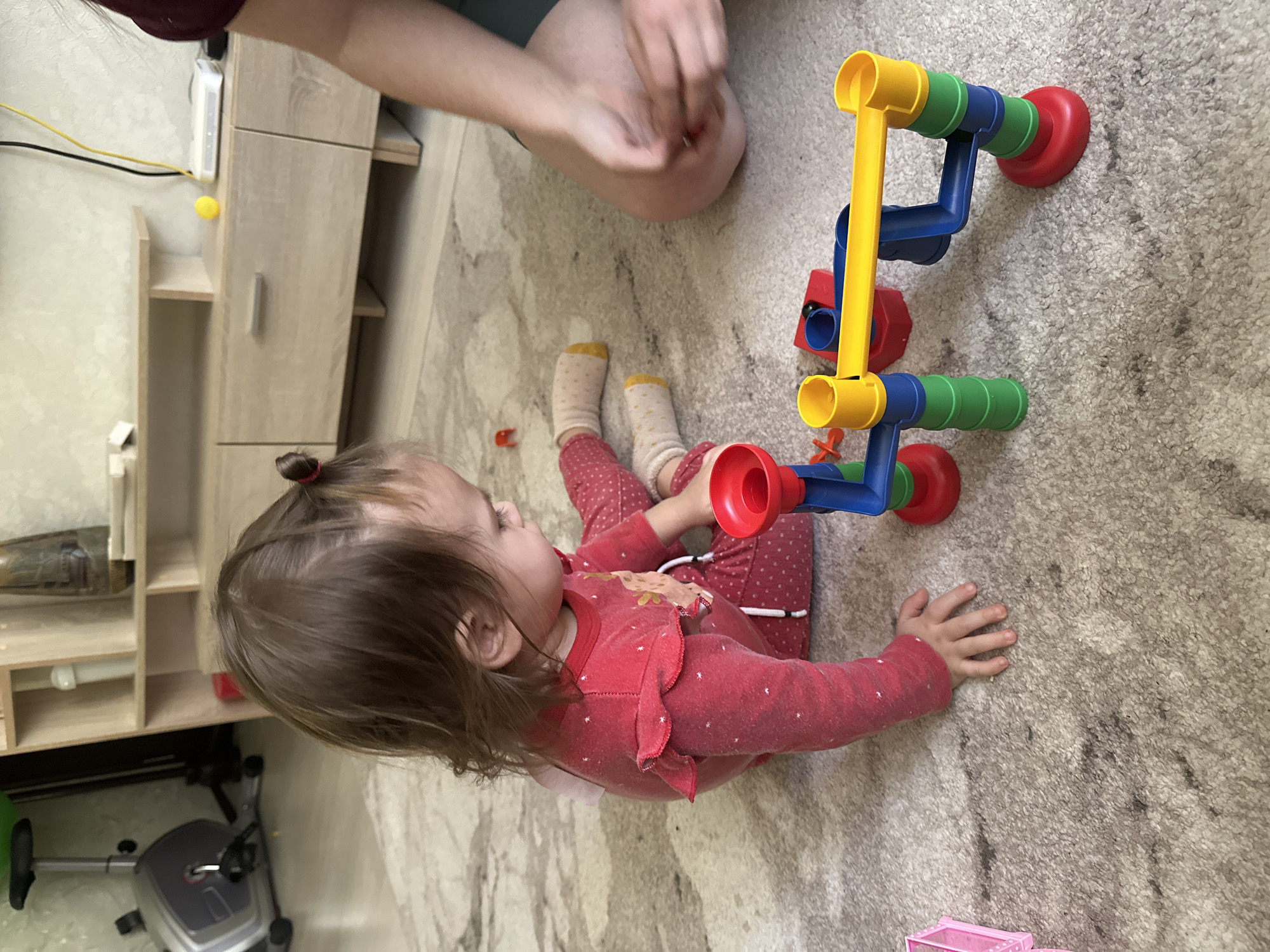 The image displays an indoor scene, likely a home in Uva, Russia, centered around a young child and toys. A toddler, appearing to be a girl with brown hair tied in a small bun, is seated on a light-colored, patterned carpet. She is dressed in a red, polka-dotted long-sleeved onesie featuring a light-colored appliqué on the chest and wears patterned socks with yellow cuffs. Her attention is focused on a brightly colored, multi-piece construction toy, possibly a marble run or similar building set, which stands on the carpet in front of her. The toy is made up of yellow, blue, green, and red plastic components.
Partially visible in the upper section of the frame are an adult's hands and lower arms, suggesting another person is also on the floor or leaning in, holding a small red object. In the background, light-toned wooden furniture, including shelving units and a cabinet, is visible. A yellow electrical cord, a white electronic device (possibly a router), and a small yellow ball are on top of one of these furniture pieces. An exercise bike is partially visible in the lower-left corner of the image, and a small pink plastic toy container can be seen on the carpet in the bottom right. Several small red plastic toy pieces are scattered near the toddler.
The scene captures a calm, daytime moment of play, emphasizing the child's engagement with the colorful toy in a typical domestic setting. There is no discernible text visible within the image.