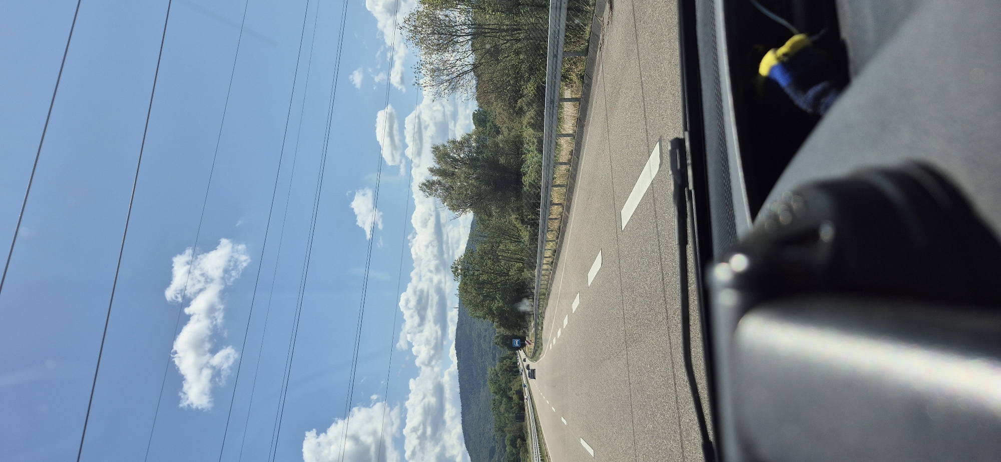 The image shows a view from inside a vehicle driving on a multi-lane highway. The highway has several lanes of traffic, marked by white lines. The sky is blue with scattered white clouds, and there are power lines crossing the sky. On the right side of the highway, there is a guardrail and a dense line of trees and bushes. In the distance, a tree-covered hill rises, with a structure, possibly a tower, visible at its summit. The foreground shows the dashboard of the vehicle, out of focus, with a portion of a dark grey interior and a blurred object with blue and yellow colors. The image appears to have been taken on a sunny day. Based on the context provided by the location Baden-Baden, Germany, this highway is likely part of the Autobahn system.