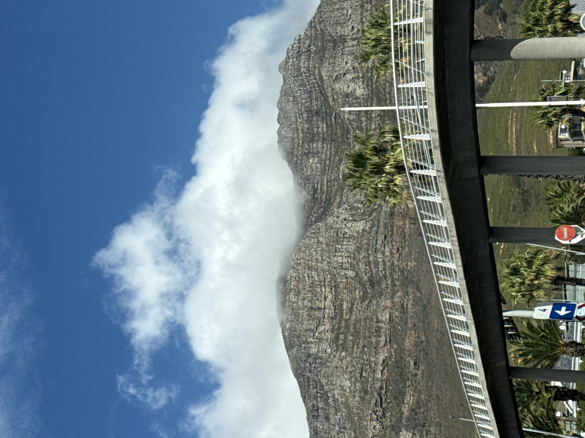 The image depicts Table Mountain in Cape Town, South Africa. The mountain's rocky face is partially obscured by a thick layer of white clouds. A road or bridge structure with a white railing runs along the right side of the image, with palm trees and traffic signs visible below. The sky is blue with scattered white clouds.