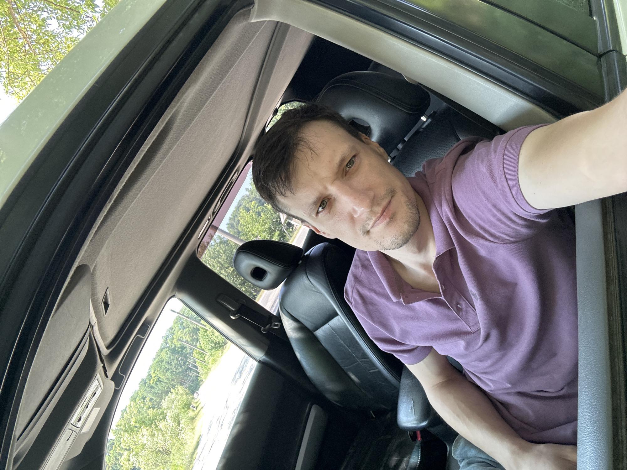 The image shows a man sitting in the driver's seat of a car, taking a selfie. He is wearing a purple polo shirt and has short brown hair and green eyes. The car appears to be a Nissan X-Trail, based on the interior details. The car's sunroof is open, and trees and a road are visible in the background. The man is looking directly at the camera with a neutral expression. The scene appears to be outdoors, possibly in a rural area given the trees and unpaved road visible outside the car.