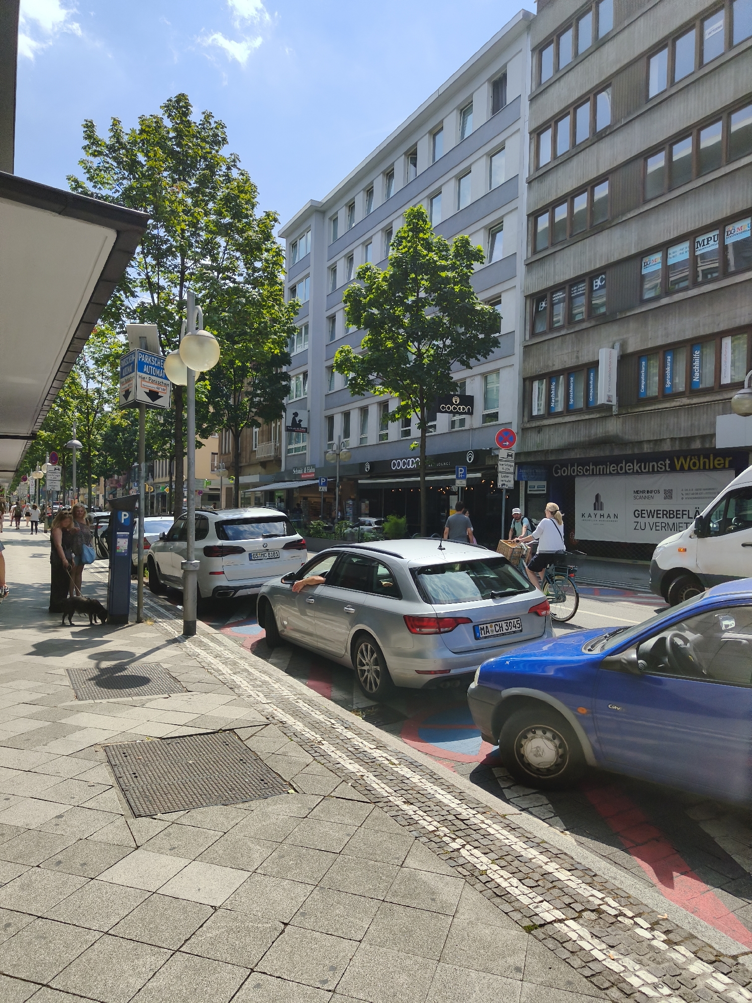 The image depicts a street scene in Mannheim, Germany, specifically on the Planken shopping street. The perspective is from the sidewalk, looking down the street on a sunny day.
In the foreground, a blue car is parked, partially obscuring the view of a silver station wagon behind it. The silver station wagon has a license plate that reads "MA CH 3945". The parking spots are marked with red and blue lines on the asphalt. A woman wearing a dark dress and a light-colored top is walking a small black dog on the sidewalk. Another woman, in a patterned dress, stands near her, looking at her phone.
The sidewalk is paved with gray square tiles and cobblestones. Several metal grates are embedded in the pavement. A lamppost with two spherical lights stands on the sidewalk, next to a parking meter labeled "PARKSCHEIN AUTOMAT" (parking ticket machine).
In the background, there are buildings lining the street, typical of a European city. The buildings have multiple stories with numerous windows. Some of the buildings house shops and businesses, with signs visible such as "Goldschmiedekunst Wöhler" and "Cocoon". Trees with lush green foliage are planted along the street, providing shade. Other vehicles, including a white van, are visible further down the street. People are walking along the sidewalk and crossing the street, indicating a bustling urban environment. The sky is blue with scattered white clouds.