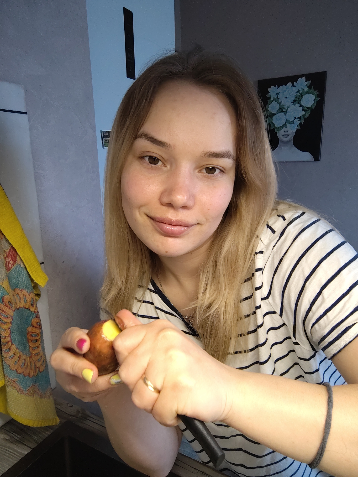 A woman is peeling a potato in a kitchen setting in Марковский, Russia. She has blonde hair, fair skin, and is wearing a striped shirt. She holds the potato in one hand and a paring knife in the other. The background includes a kitchen sink, a yellow and patterned towel, wall tiles, and a painting of a woman with flowers on her head.