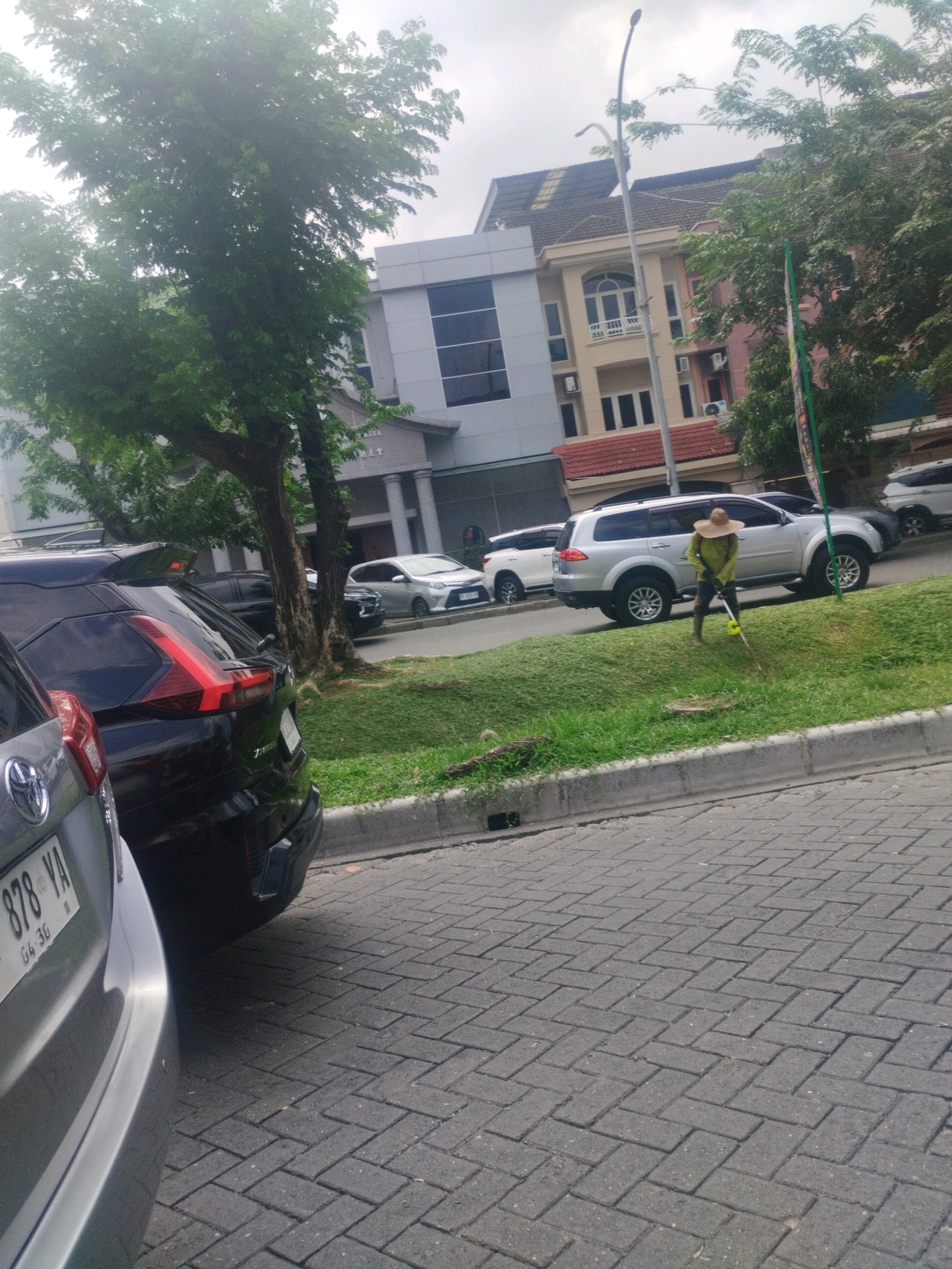 In Percut Sei Tuan, Indonesia, a man wearing a wide-brimmed hat and a light-colored long-sleeved shirt is mowing a grassy area next to a paved road. He is using a yellow-handled string trimmer. Behind him, a silver SUV is parked on the grass, and further back, several other vehicles are visible, including a black SUV in the foreground. The scene is in front of a multi-story building with a modern gray facade and a more traditional building with cream-colored walls and a pink facade. Trees with green foliage are present throughout the scene. The road is paved with gray interlocking bricks.