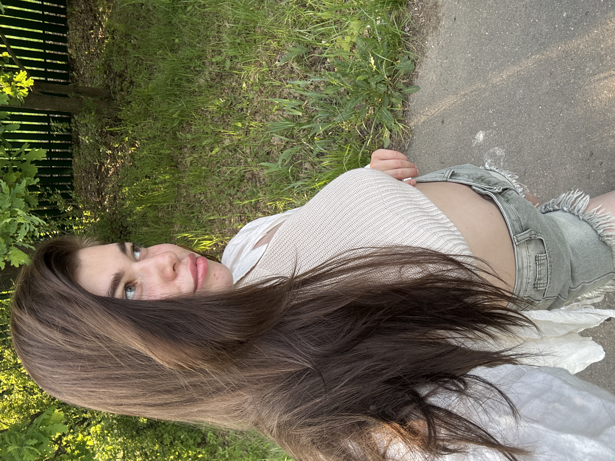 A young woman with long brown hair and blue eyes is posing outdoors. She is wearing a light-colored knit top and denim shorts. The location appears to be a park or green area with trees, grass, and a paved path. The specific location of Мещёры, Russia cannot be determined from the given image.