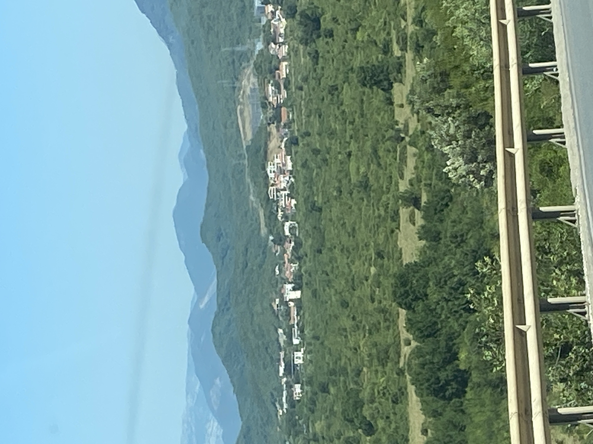 The image depicts a view of Prijevor, Montenegro, characterized by lush green hills dotted with a cluster of houses. A bright blue sky stretches across the top of the frame, with a hazy, distant mountain range visible in the background. In the foreground, a beige guardrail runs horizontally across the bottom of the image, separating the viewer from the landscape. The houses in Prijevor are small and appear to be a mix of white and terracotta-roofed structures, nestled amongst the dense foliage. The overall scene suggests a rural or semi-rural setting with a tranquil, natural environment.
