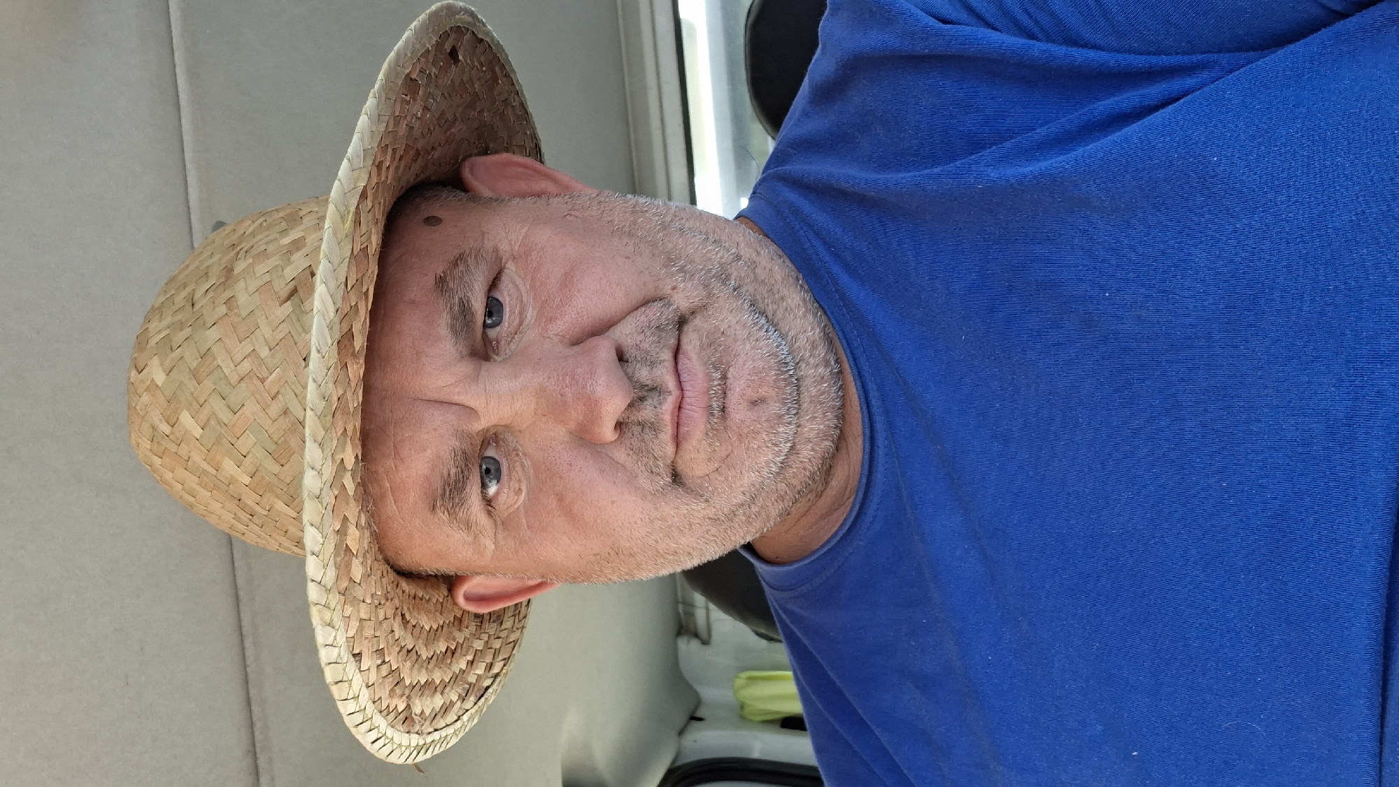 A middle-aged man wearing a straw hat and a blue t-shirt is captured in a close-up, frontal photograph. The man has a weathered face with blue eyes, and a visible stubble beard. His expression is neutral, and he appears to be indoors, possibly in a vehicle, suggested by the textured ceiling in the background. The lighting is bright, illuminating the man's features and the weave of his hat. The image is not specific to any location within the city of Hîncești, Moldova, as no distinguishing landmarks or contextual elements are present.