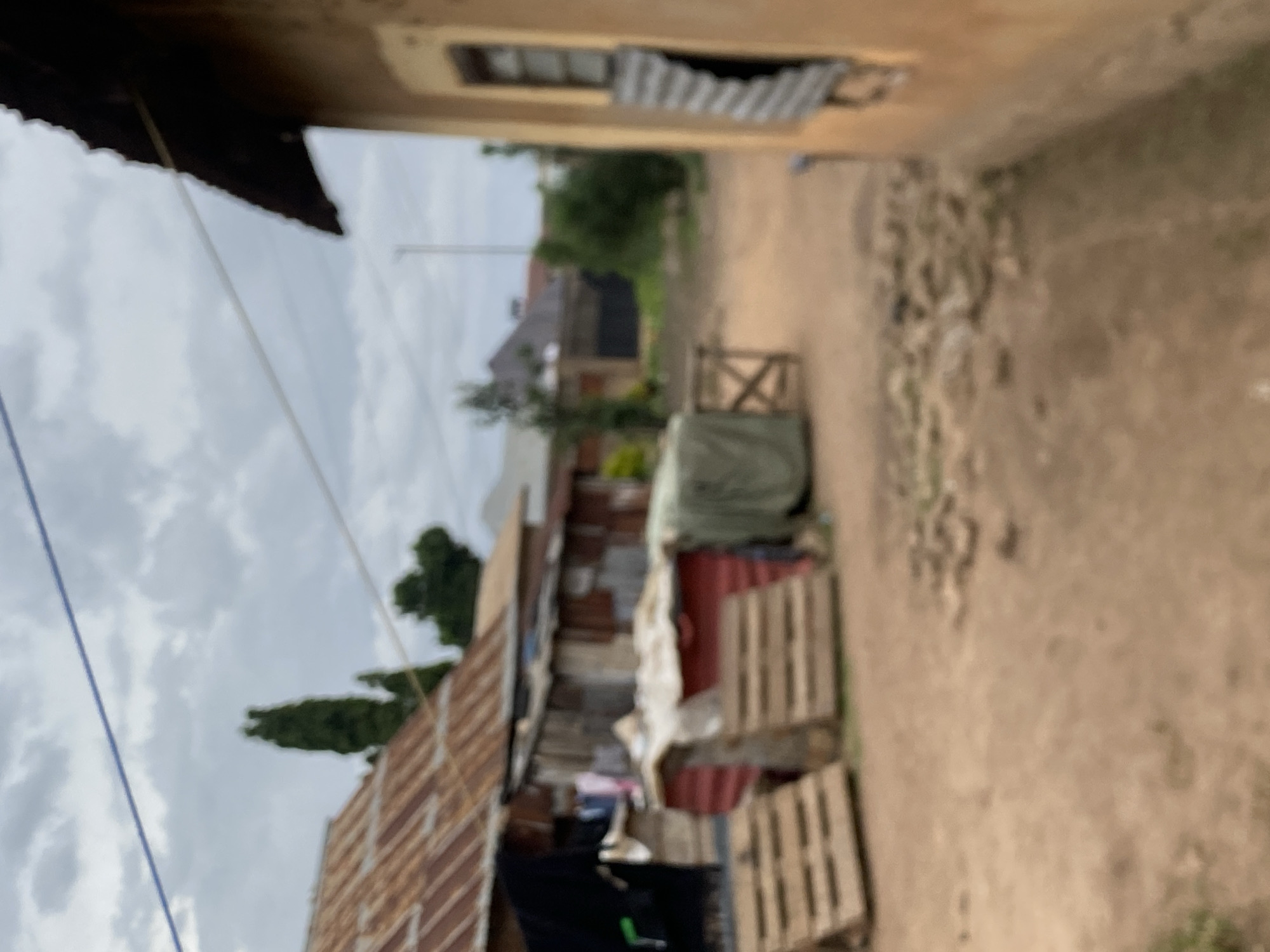 This photograph depicts an alleyway in Bukuru, Nigeria. The alleyway is unpaved. On the left, there are structures made of corrugated metal and other materials, and on the right, there is a wall or building. In the alleyway, there are stacks of wooden pallets and an object covered by a green tarp. The sky is overcast.