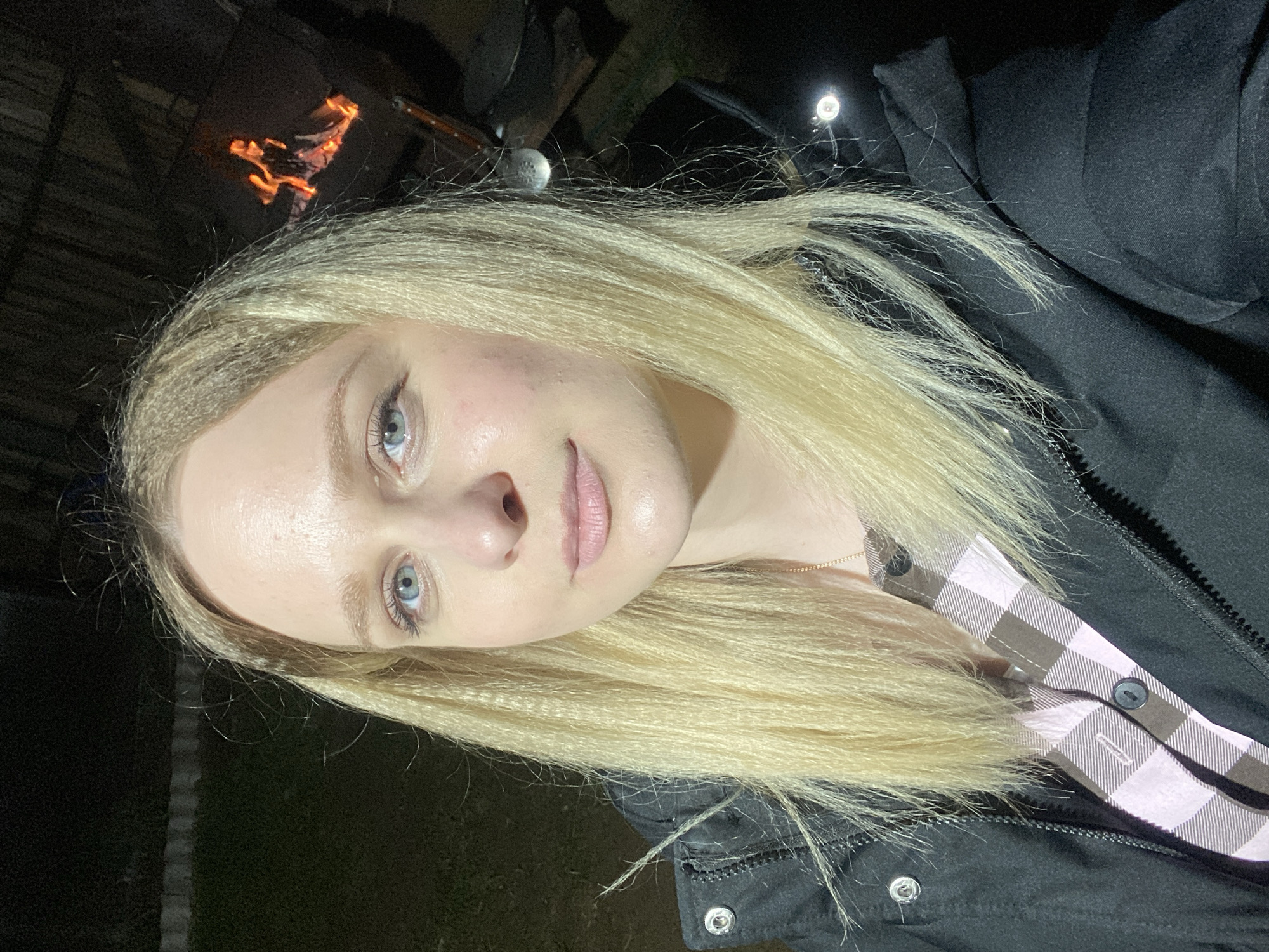 A close-up, vertical shot captures a young woman outdoors at night, likely in Kauchuk Stantsiya, Russia. She is positioned for a selfie or headshot, with her head slightly tilted. Her long, blonde hair, styled with a crimped or wavy texture, frames her face. She has light blue eyes and a neutral to slightly smiling expression, with light pink lips. Her skin appears fair.
She is dressed in a black jacket, unzipped to reveal a pink and brown plaid button-up shirt underneath. A thin gold chain necklace is visible around her neck. In the dark background, to the upper left, a small fire or campfire with visible flames glows. Further back and to the right, a bright, small light source, possibly a headlamp or lantern, is visible. Another small, light-colored object, resembling a golf ball on a stick, can be seen near her hair on the right side of the frame, possibly part of a decorative item or a light fixture. The general setting is dark, suggesting nighttime, with hints of grass or ground visible on the left side. No visible text is present in the image.