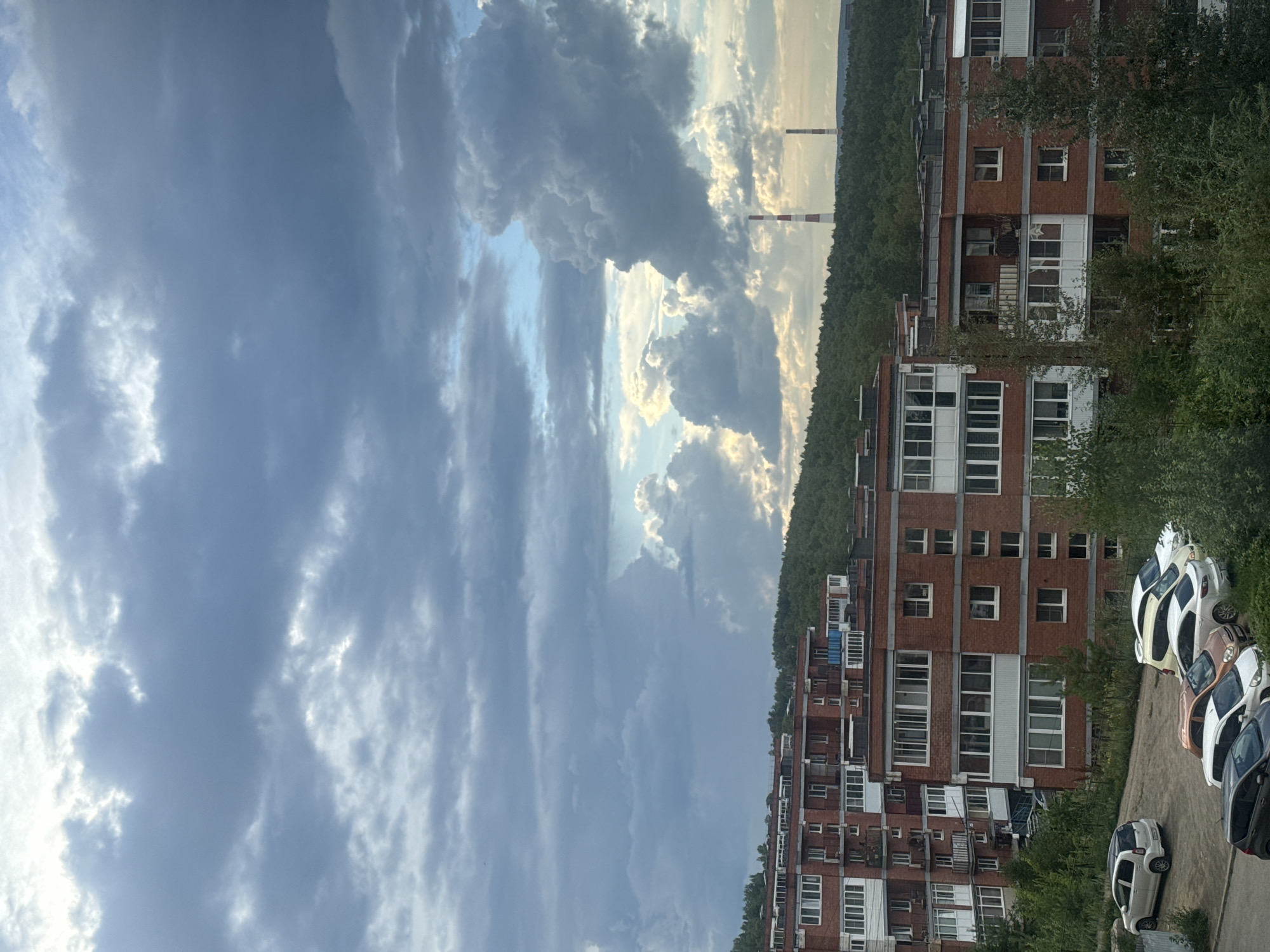 This is an outdoor, eye-level, medium shot of a residential area with apartment buildings and a cloudy sky. The apartment buildings are made of red brick and have white balconies and window frames. Several cars are parked in front of the buildings. The sky is filled with dramatic, dark clouds, with a sliver of blue sky and sunlight peeking through in the distance. Two smokestacks, one red and white striped and the other plain red, are visible in the far distance. The scene is set in the city of Markova, Russia.