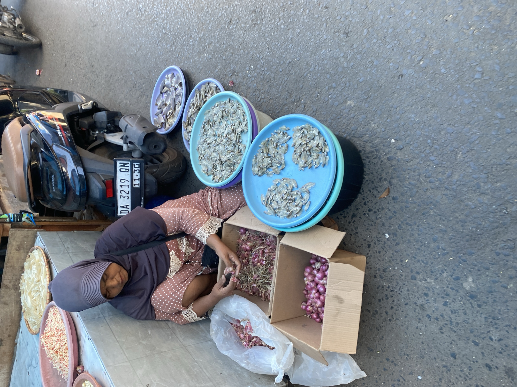 A woman wearing a dark purple hijab and a brown and white polka dot dress is seated on the ground next to a market stall in Desa Tunggul Irang, Indonesia. She is sorting through dried anchovies and red onions. In front of her are two cardboard boxes filled with red onions, a plastic bag also containing red onions, and several shallow blue and purple bowls filled with dried anchovies. Behind her, a dark-colored motorcycle is parked. The ground is asphalt.