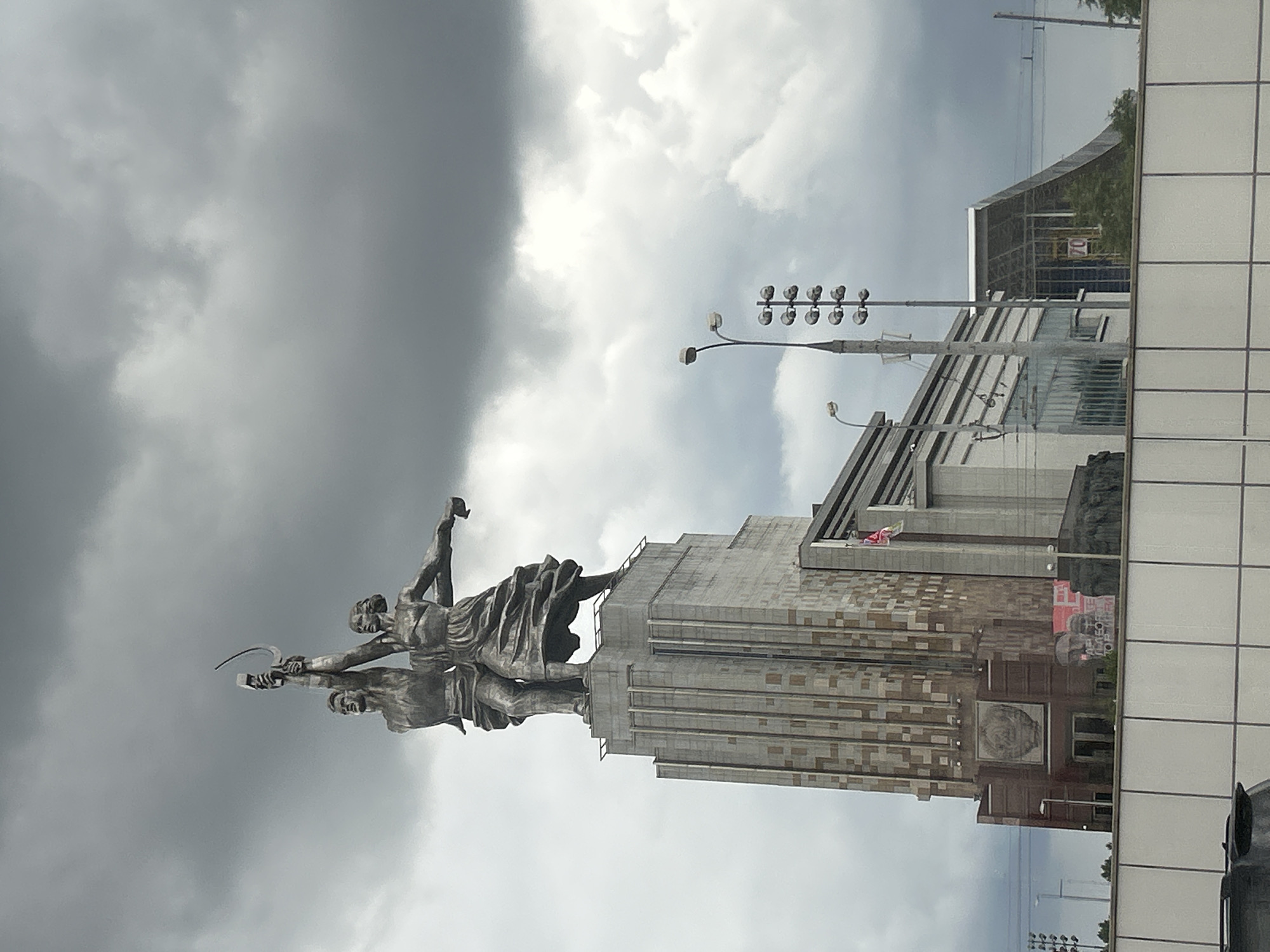 The image shows a statue of a man and a woman holding a sickle and hammer, symbolizing the worker and the collective farm worker, atop a tall, art deco-style building. This iconic sculpture, "Worker and Kolkhoz Woman," is a prominent landmark in Moscow, Russia. The statue is made of stainless steel and stands at 24.5 meters (80 feet) tall. The building it rests on is part of the VDNKh (Exhibition of Achievements of National Economy) complex, a vast public park and exhibition center in Moscow.
The sky is overcast with dark, dramatic clouds, suggesting a stormy or moody atmosphere. The lighting is diffused, casting a shadowless and somewhat muted tone over the scene. In the foreground, there is a white wall, possibly a barrier or part of a lower structure, with a glimpse of a car roof on the left. Beyond the central building, other structures of the VDNKh complex are visible, including modern buildings with glass facades and a large, sloping roof structure. Streetlights and electrical wires are also present, indicating an urban environment. The overall impression is one of grandeur and imposing scale, with the statue and the architecture creating a powerful visual statement against the dramatic sky.