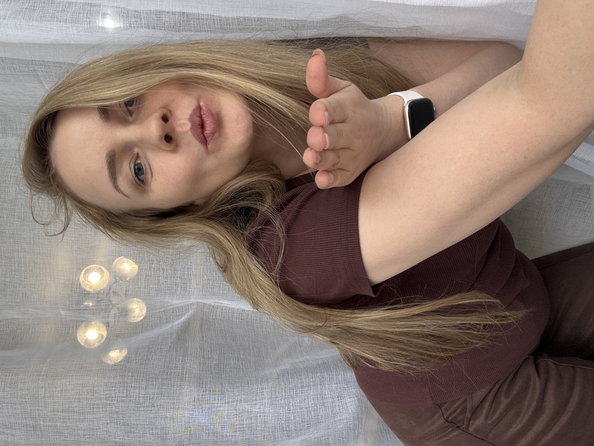 A blonde woman is posing indoors. She is wearing a maroon top and pants, and has a white smartwatch on her left wrist. She is holding her right hand up, slightly extended towards the camera. There is a sheer white curtain behind her, and a reflection of a light fixture is visible through the curtain. The location is unknown beyond the statement that the image was taken in Chaikovsky, Russia.