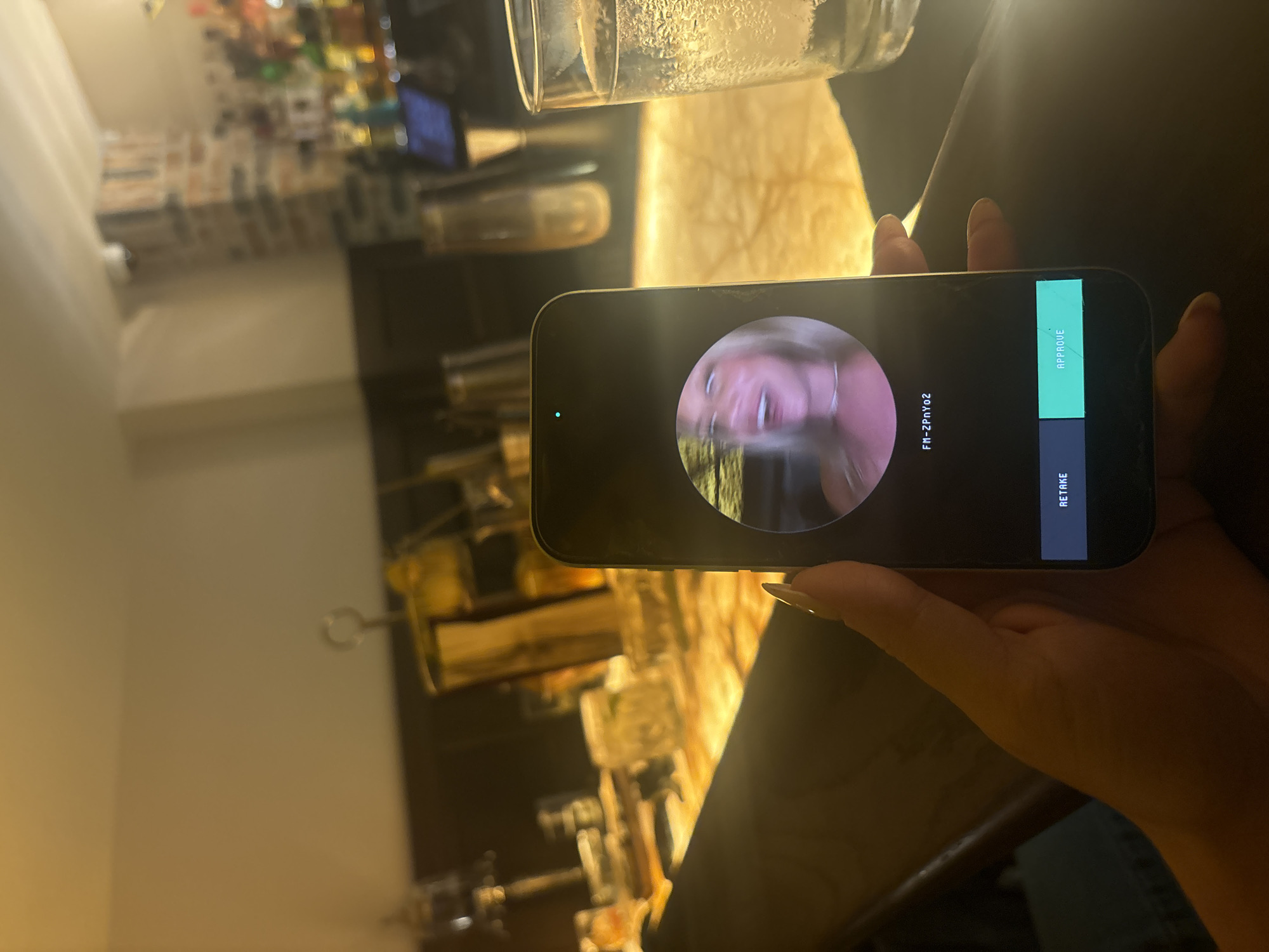 The image captures a first-person perspective of a hand holding a smartphone in a dimly lit bar or restaurant setting in Miami, United States of America.
The primary subject is a right hand with long, light-colored manicured fingernails, holding a dark-colored smartphone vertically. The phone screen displays an interface for reviewing a captured image or video. A circular frame in the center of the screen shows a close-up of a blonde woman with a wide-open mouth, appearing to be mid-laugh or speaking, her eyes wide, and wearing a light-colored necklace. The image of the woman is slightly blurred, suggesting motion. Below the circular frame, the alphanumeric code "FM-2PnYo2" is displayed. At the bottom of the screen, two rectangular buttons are visible: "RETAKE" on the left (dark grey) and "APPROVE" on the right (teal/green). A small, green light indicator is present in the top-left area of the phone screen, near the dynamic island/notch.
The background is out of focus, indicating a shallow depth of field. To the right of the phone, a clear glass with ice and a beverage, likely water, shows condensation on its exterior. The surface beneath the phone and glass is dark and reflective, possibly a bar counter. The wider background reveals a warm, ambient environment characteristic of a bar, with various blurred bottles and glasses on shelves or behind the counter. A brick wall or pillar is visible in the mid-right background, along with other indistinct decor. The lighting is predominantly warm and creates reflections on surfaces. A light-colored ceiling is visible at the top, and a small, white, dome-shaped object (possibly a security camera) is faintly discernable near the ceiling on the right.