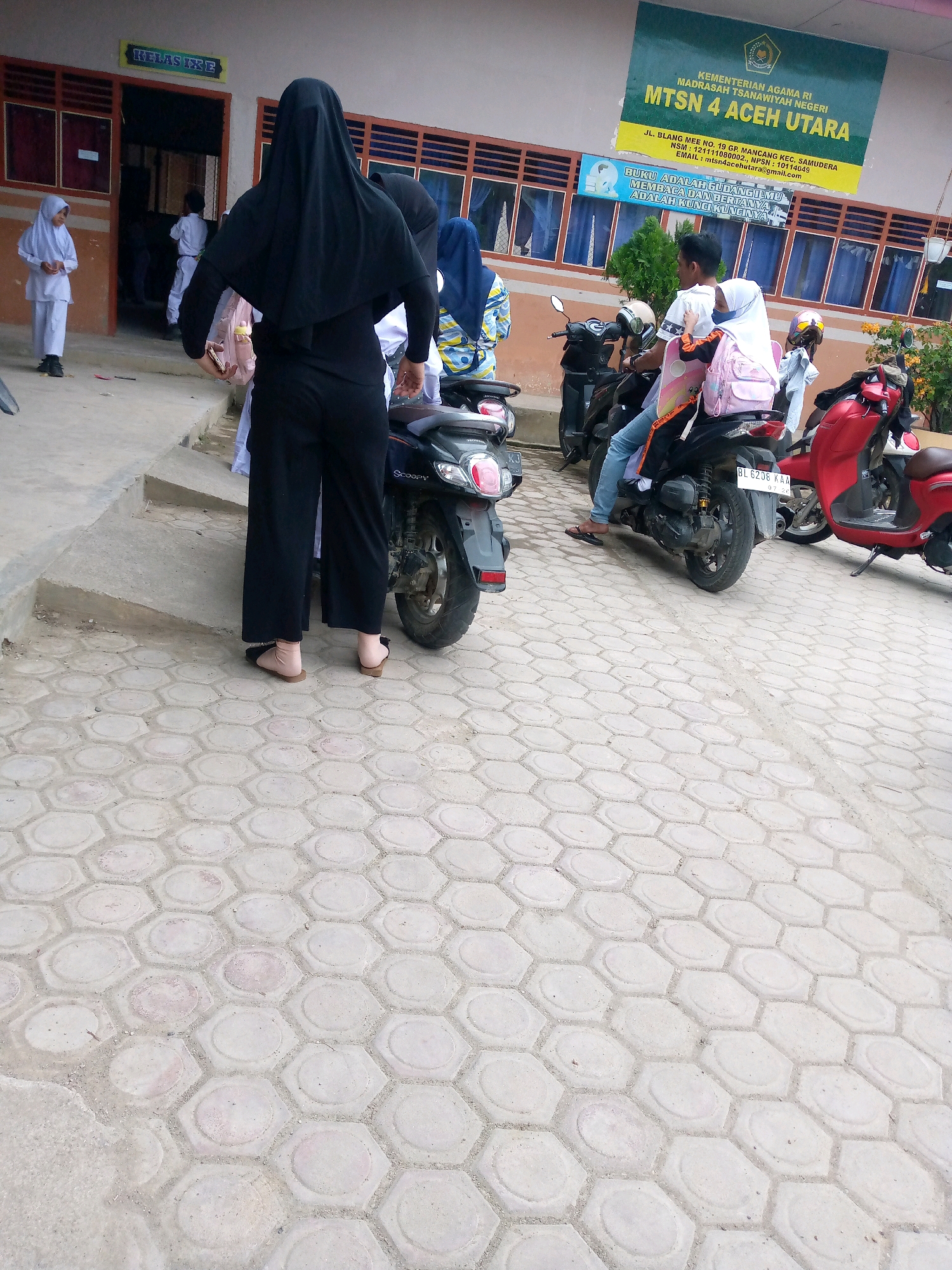 The image depicts the outdoor area of a school in Mesjid, Indonesia. In the foreground, a person wearing a black hijab and black clothing stands with their back to the camera, holding a pink backpack. To the left, a young girl in a white hijab and uniform stands on a step. In the middle ground, two people are seated on a scooter; one person is wearing a white shirt and jeans, and the other, a girl in a white hijab, is also on the scooter. Further to the right, a red scooter is parked. The school building is visible in the background, with signs indicating "KELAS IX E" and a larger sign for "KEMENTERIAN AGAMA RI MADRASAH TSANAWIYAH NEGERI MTSN 4 ACEH UTARA". The ground is paved with hexagonal concrete tiles.