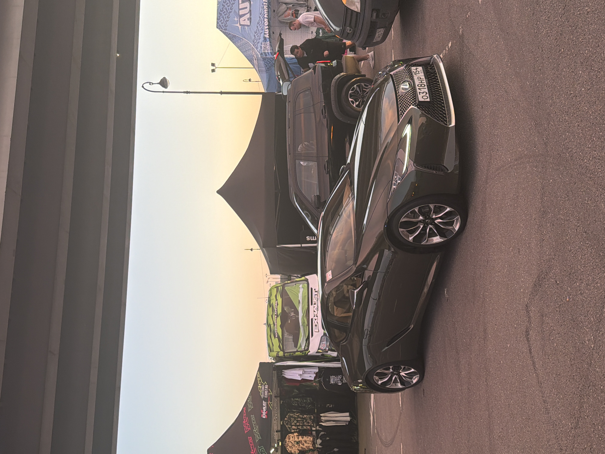 An outdoor event is taking place under a concrete overpass. In the foreground, a dark green Lexus LC 500 is parked facing right. The car has a license plate reading "0318HP 154". To the left of the Lexus, a tent with apparel for sale is set up, featuring racks of clothing. Behind this tent, a white van with green camouflage patterns is parked, also displaying "EXHAUST" branding.
In the background, a black SUV is parked, with its rear hatch open, and two men are standing near it. Another tent, this one blue and white, is visible to the right, with the word "AUTO" and "AUTOFAMILY" visible on it. A street lamp with a decorative design stands in the middle ground. The sky is a gradient of pale orange and blue, indicating either sunrise or sunset. The ground is paved asphalt, with some tire marks visible.