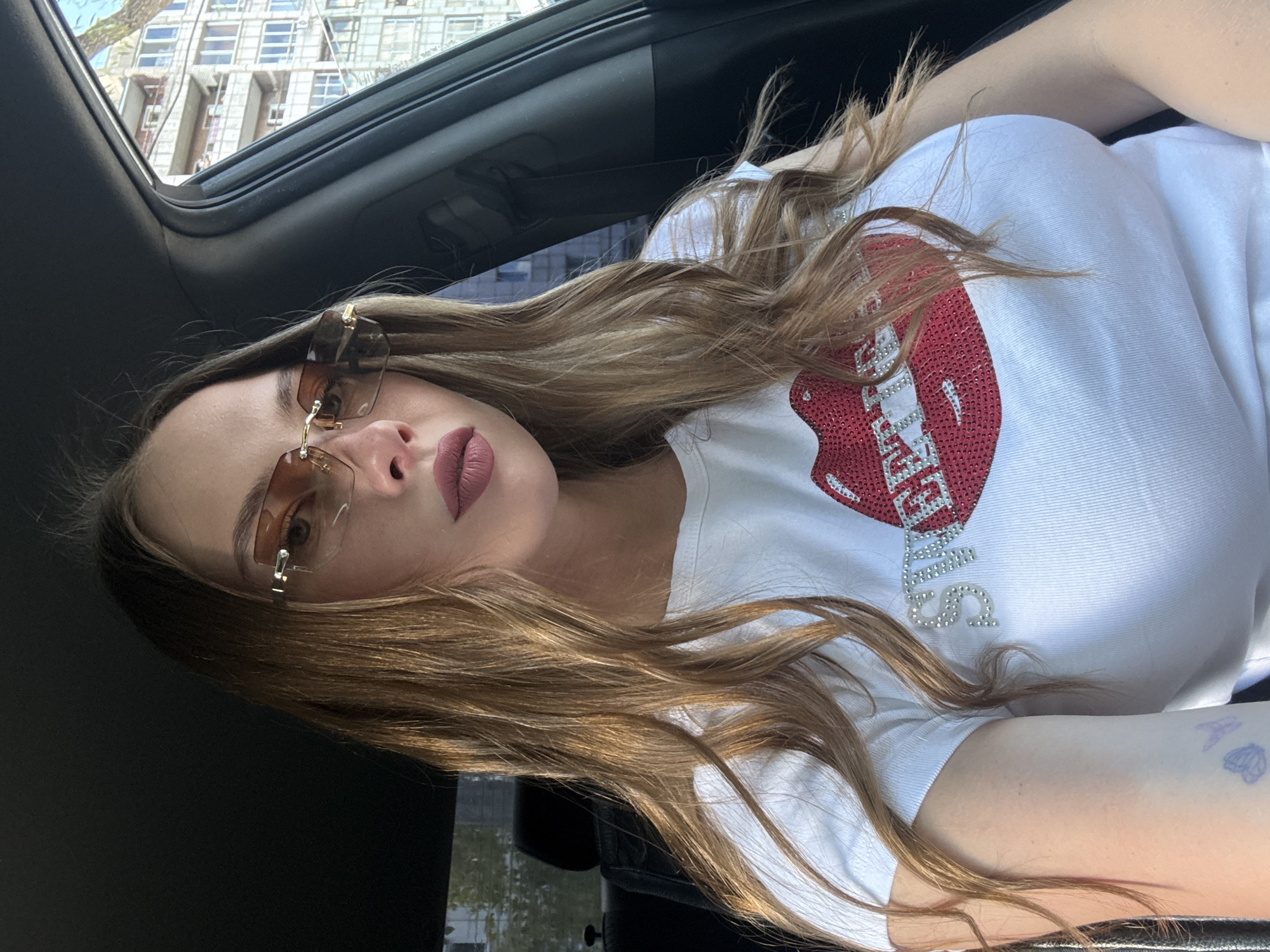 A young woman with long, wavy brown hair and light skin is sitting in the driver's seat of a car. She is wearing rimless, brown-tinted sunglasses and a white t-shirt with a large red sequined lip design and the word "SWEETIES" written in rhinestones. Her lips are painted a matte pink color. She has a small butterfly tattoo on her left arm. The car's interior is visible, including a black headrest and part of the dashboard. Outside the car, a multi-story residential building with balconies can be seen, suggesting an urban environment. The overall lighting indicates it is daytime. The building visible in the background is consistent with typical residential architecture found in Perm, Russia, specifically in areas with mid-rise apartment complexes.