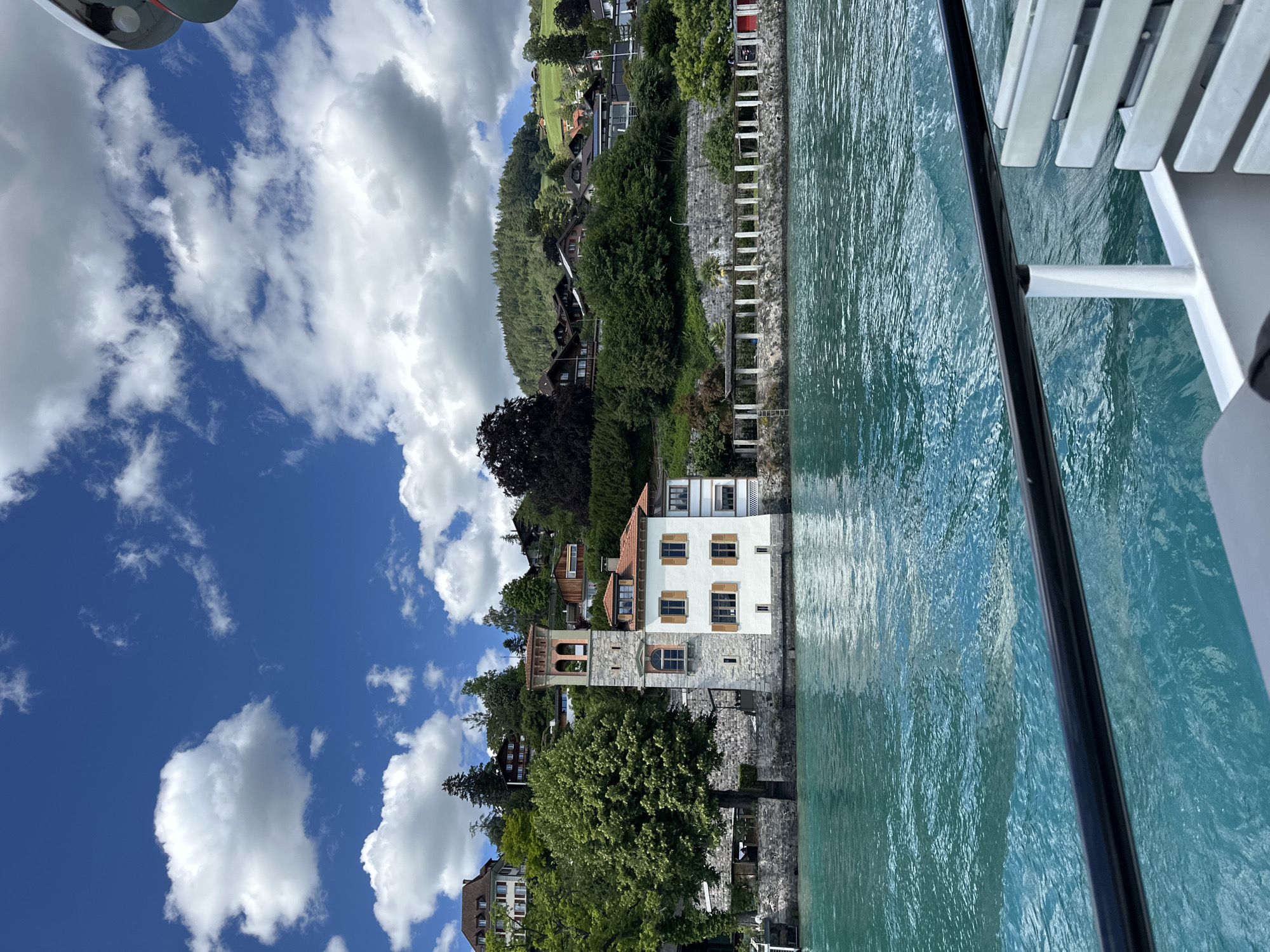 This is a full shot of the picturesque town of Oberhofen am Thunersee, Switzerland, captured from the perspective of a boat on the turquoise waters of Lake Thun. The scene unfolds under a bright blue sky dotted with fluffy white clouds.
Dominating the left side of the frame is a striking white building with a prominent stone tower, featuring arched openings and a terracotta roof. This building appears to be situated on the lakeshore, with a stone wall and lush greenery surrounding it. Several other houses and structures are visible further up the hillside, nestled amongst dense trees and vegetation, suggesting a residential area on the slopes. The architecture of the houses is varied, with some featuring traditional wooden elements.
The water in the foreground is a vibrant blue-green, reflecting the sky and the surrounding scenery. Ripples on the water's surface indicate gentle movement. The edge of the boat on which the photograph is taken is visible in the lower right corner, with a black railing and white seating.
The overall impression is one of serene natural beauty and charming lakeside architecture. The image captures a quintessential Swiss lakeside town on a clear, sunny day.