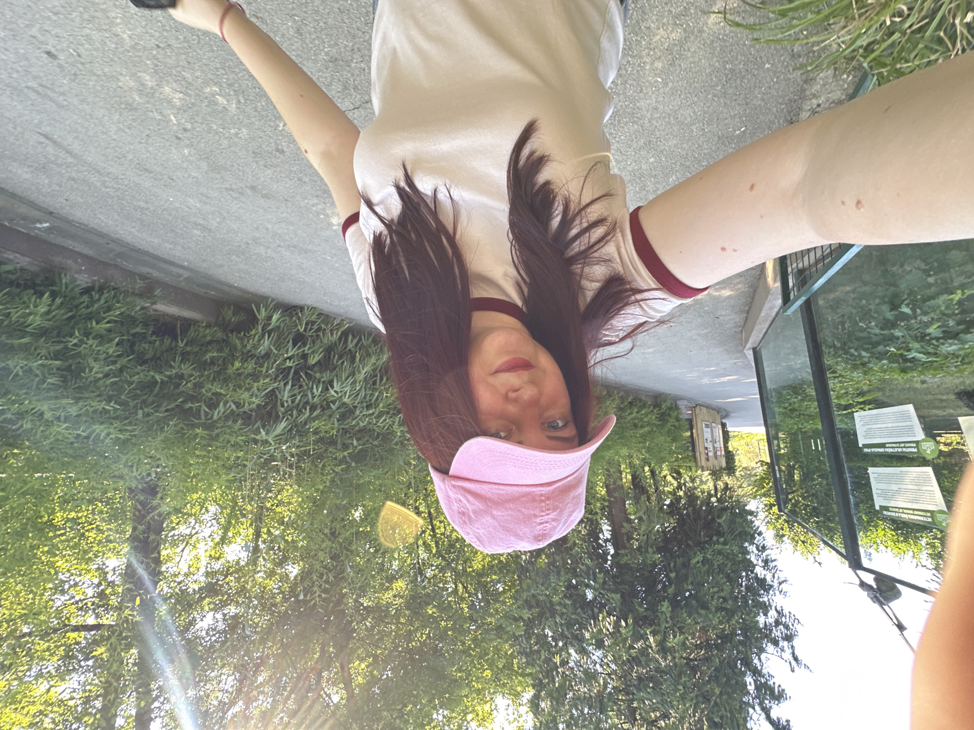 A young woman wearing a pink baseball cap and a white t-shirt with burgundy trim is taking a selfie outdoors. She is walking on a paved path next to a glass enclosure with informational signs. Lush green trees and foliage surround the path. The sun is shining brightly, casting lens flares and dappling the scene with light. The environment appears to be a zoo or park, likely within the City of Zagreb, Croatia, given the text on the signs.